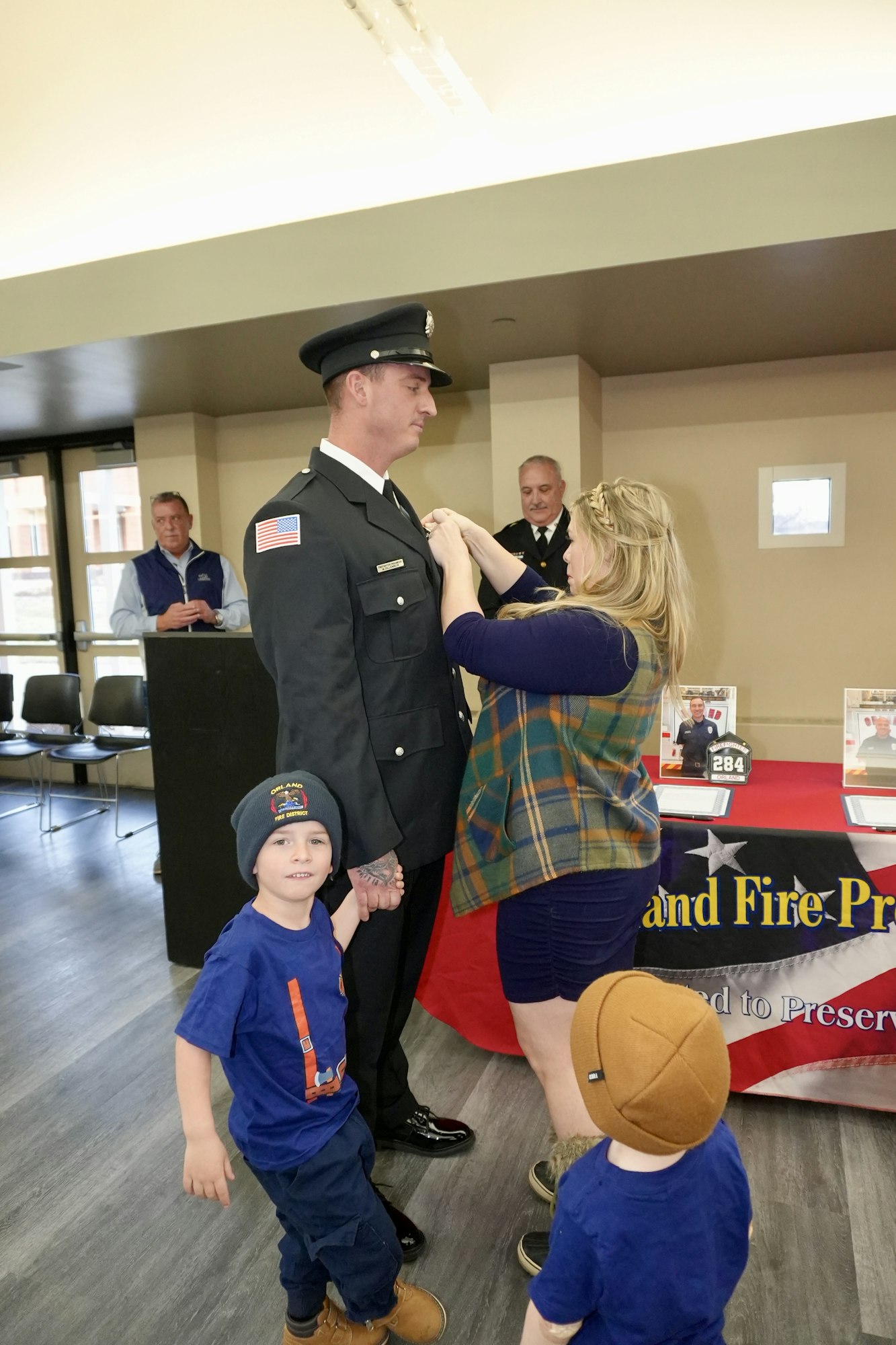 A woman pins a badge on a firefighter in uniform during a ceremony, with two children nearby and onlookers in the background.
