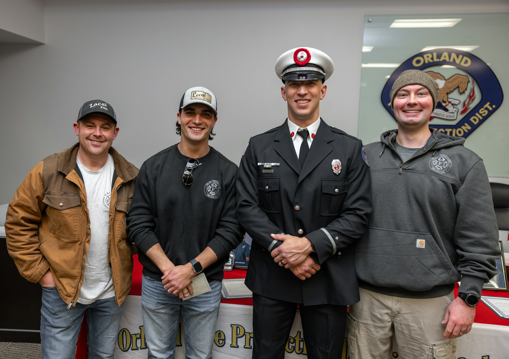 A group of four men, including one in a firefighter uniform, pose together in an office setting behind a protection district sign.