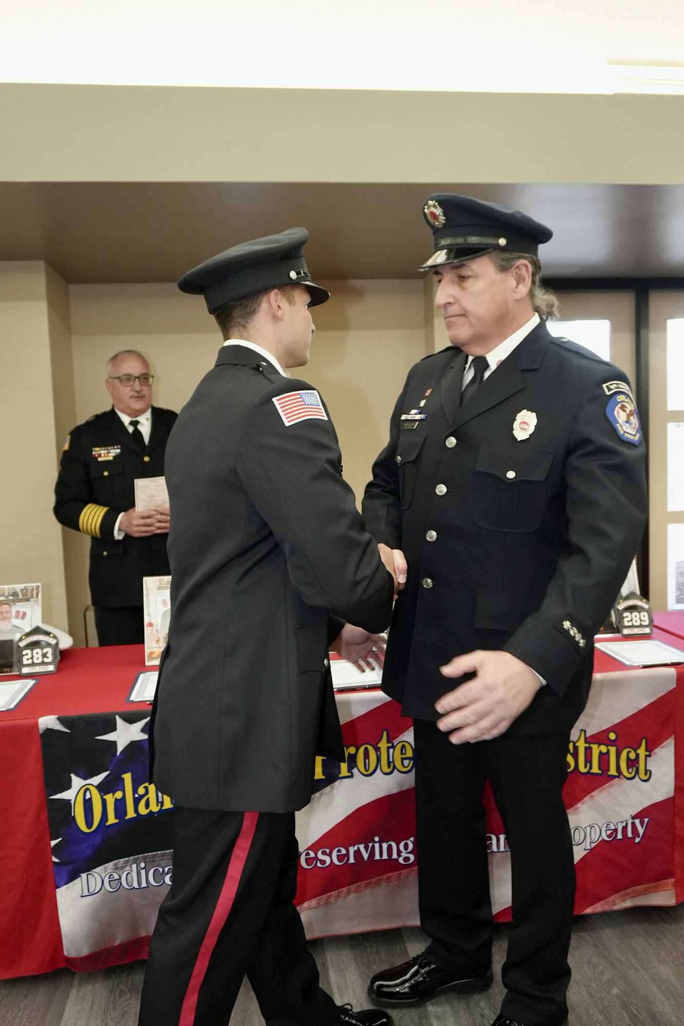 Two uniformed firefighters shaking hands, another in the background, in front of a table with a banner.