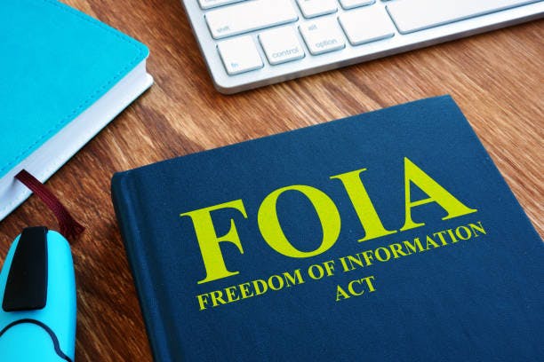 The image features a book titled "FOIA: Freedom of Information Act," with a pen and notebook on a desk, beside a keyboard.