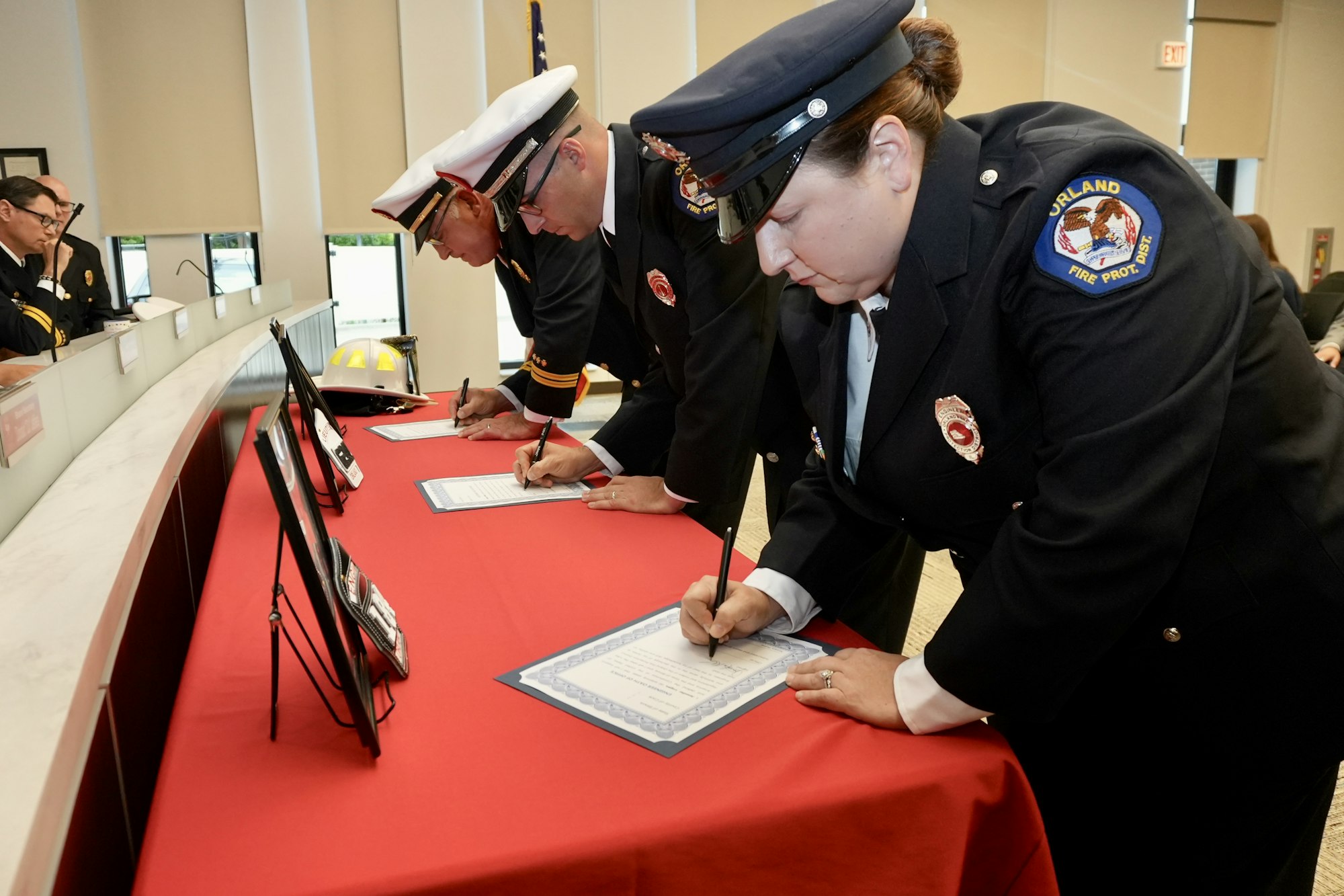 Firefighters in uniform signing documents at a red-draped table.