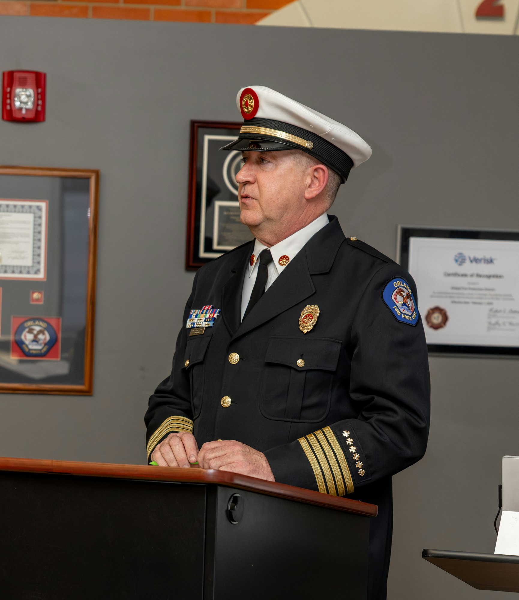 The image shows a man in a military-style uniform with a hat, speaking at a podium in front of framed certificates and awards.
