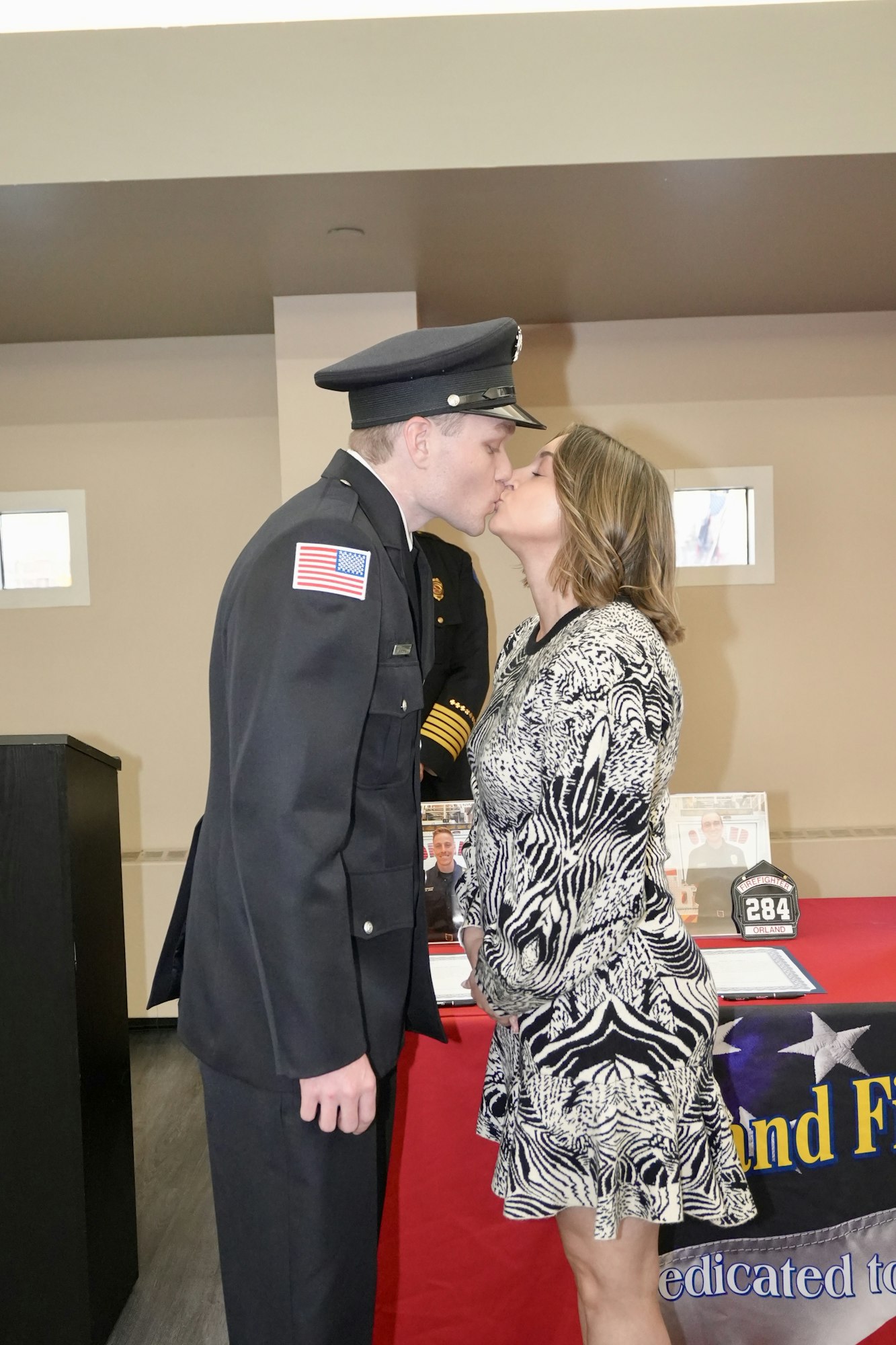 A person in a uniform and a woman wearing a patterned dress share a kiss by a red table with a badge display.