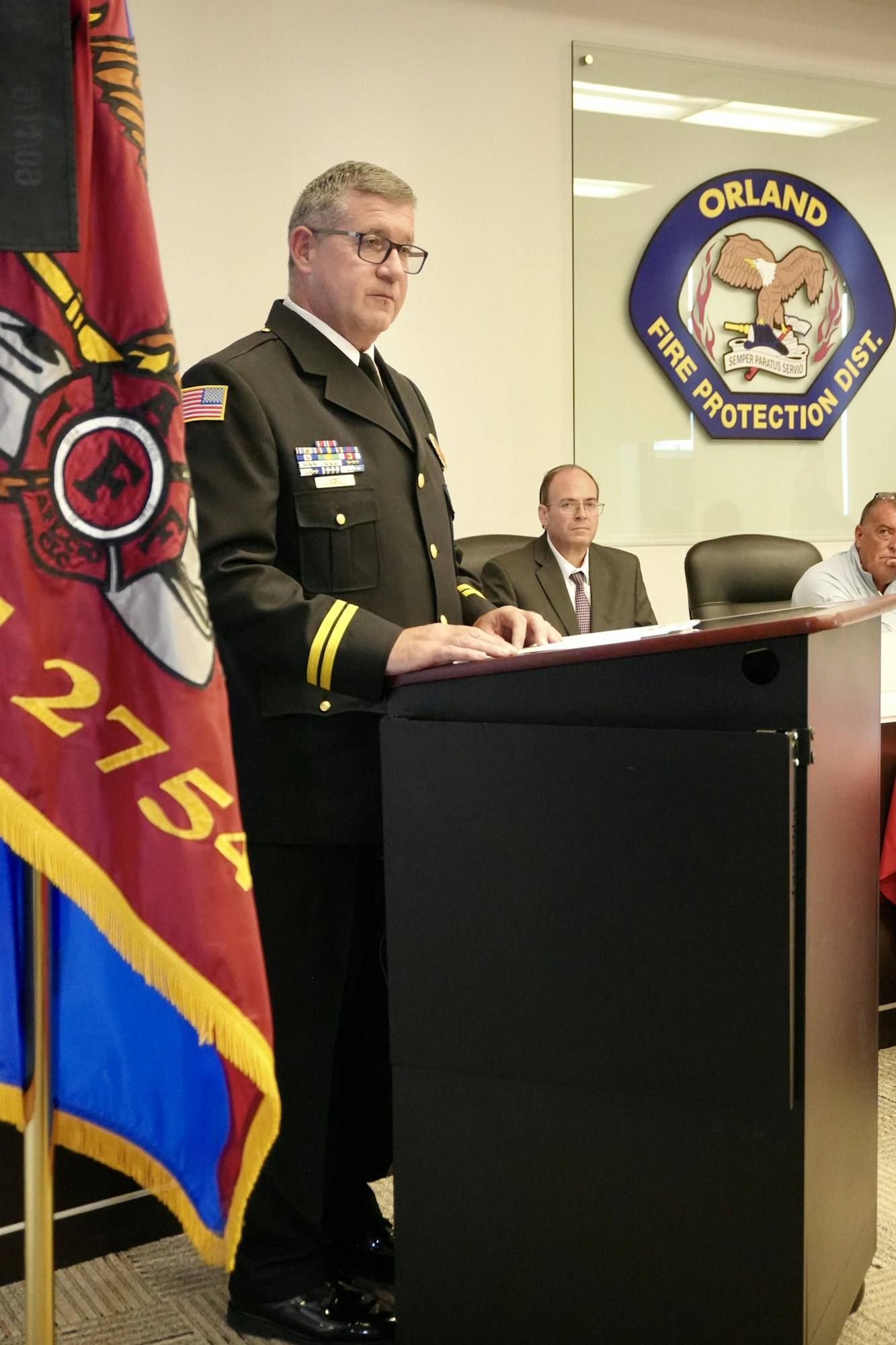 A person in a uniform speaks at a podium with a "Fire Protection Dist." emblem on the wall behind. A flag and two seated individuals are visible.