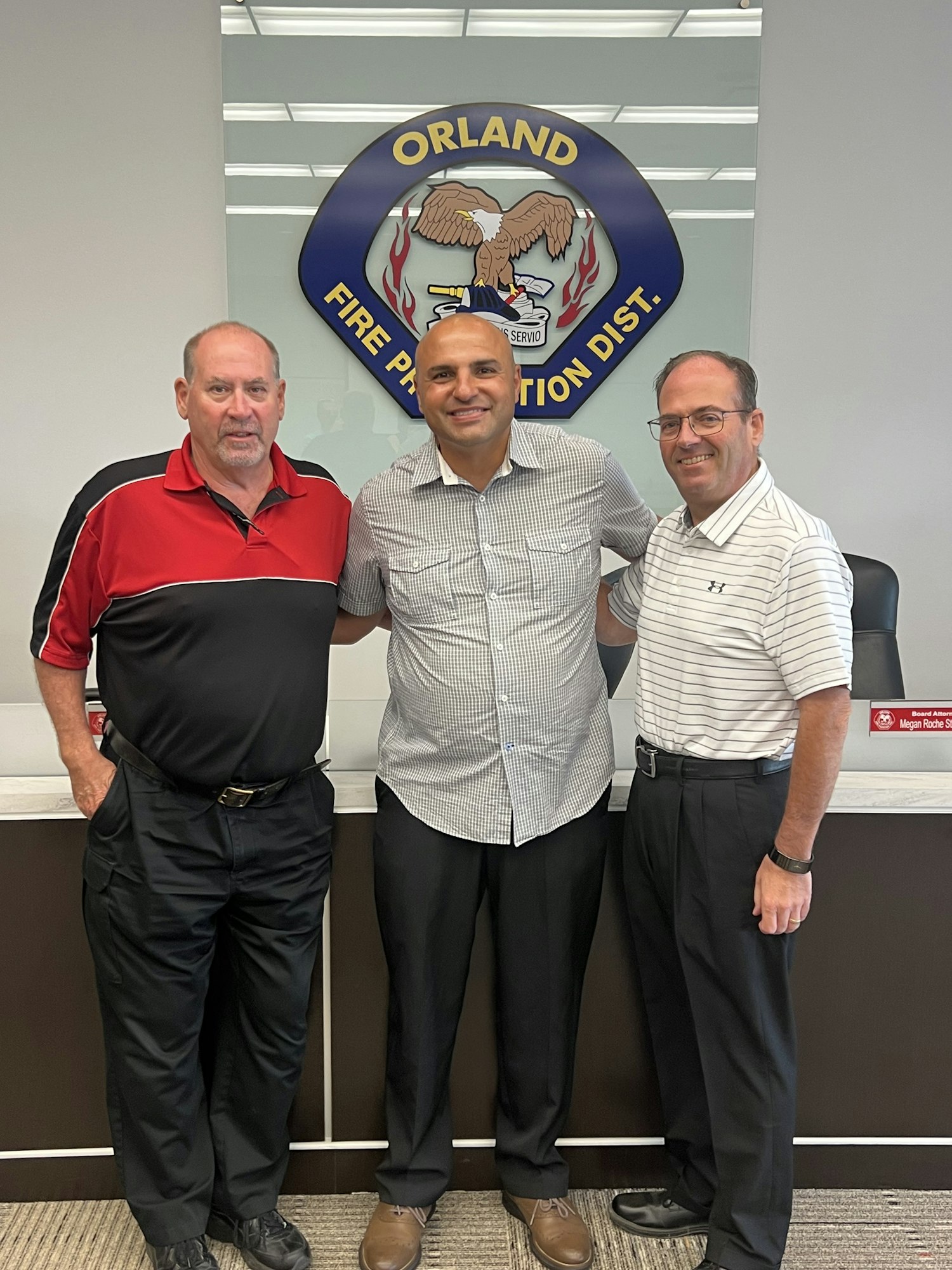 Three men standing in front of a wall with the Orland Fire Protection District logo.