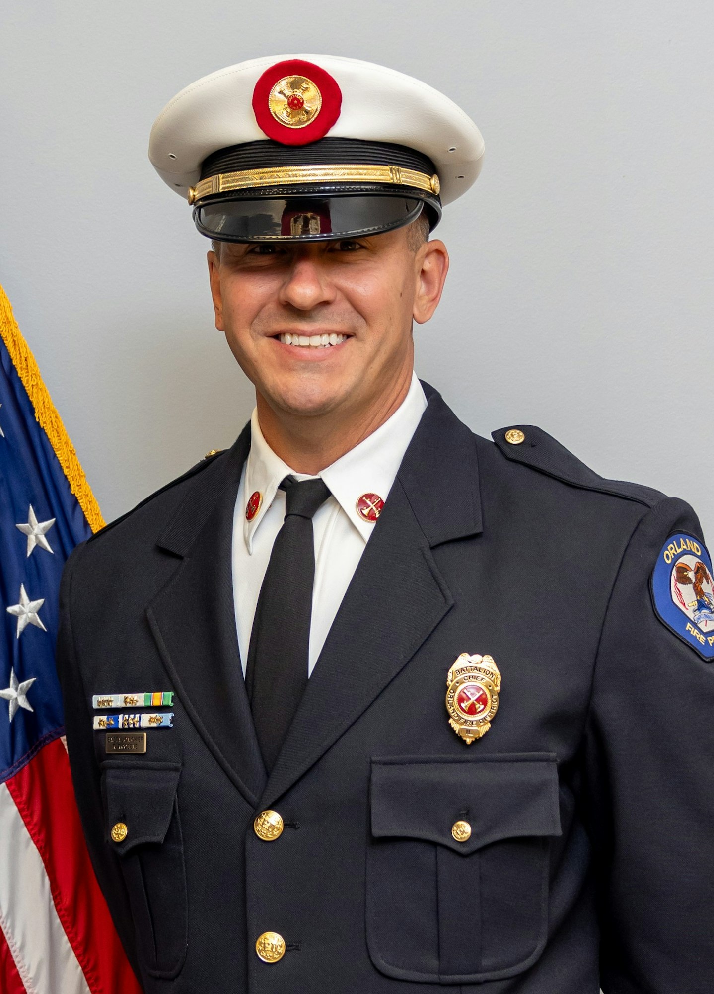A smiling individual in a uniform, likely a firefighter or fire chief, standing in front of an American flag backdrop.