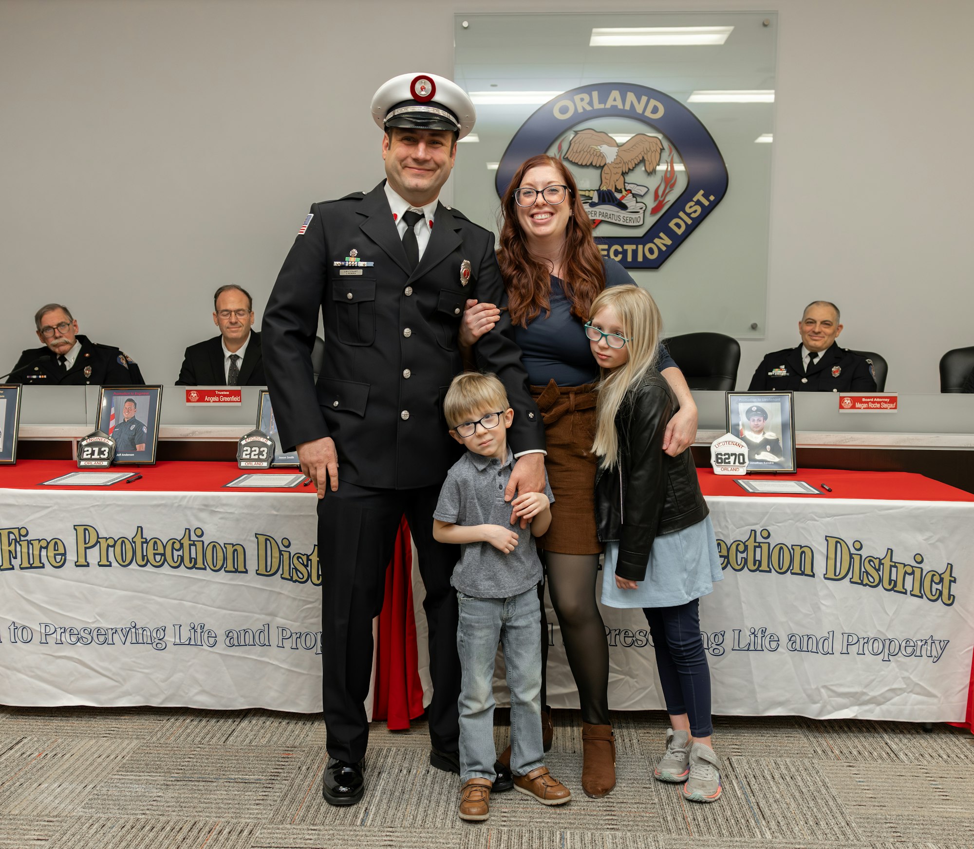 A firefighter in uniform poses with his family at a ceremony, with officials seated behind him and a banner promoting fire safety.