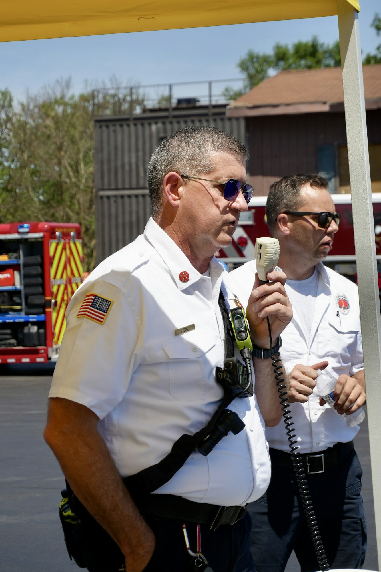 Two men in white uniforms, one speaking into a microphone, with emergency vehicles in the background.