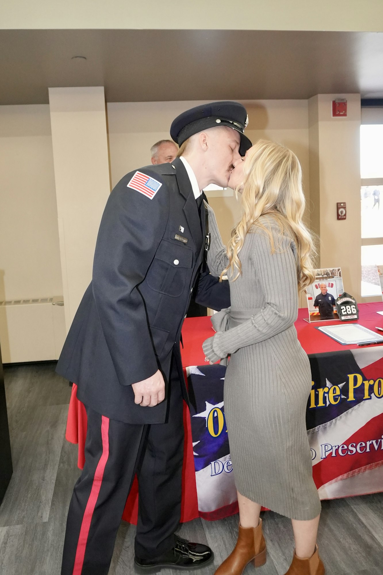 A person in a uniform kisses another person in a gray dress near a table with a flag-themed cover.