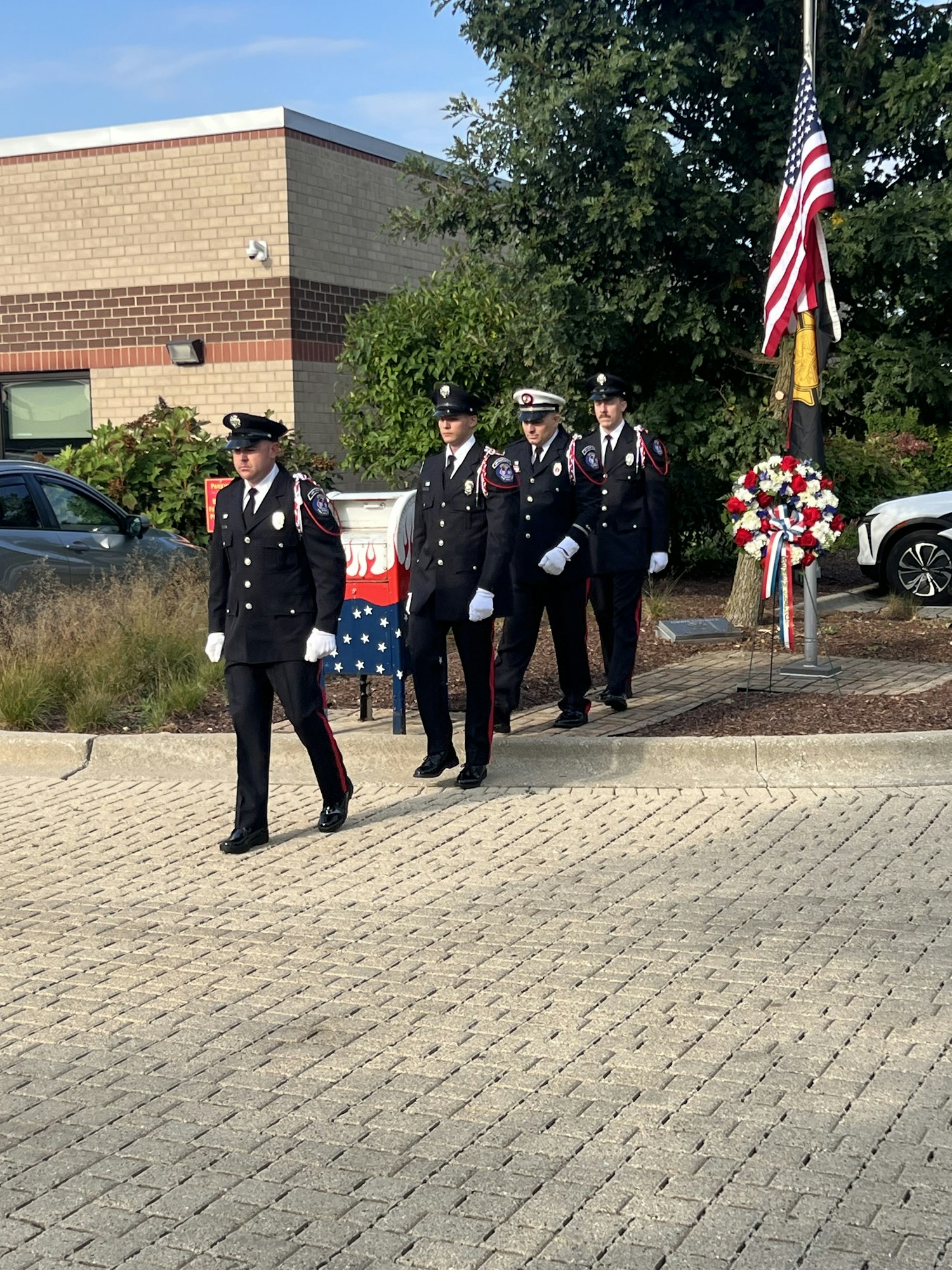 Uniformed individuals in a ceremony by a flagpole with the US flag and a patriotic wreath.