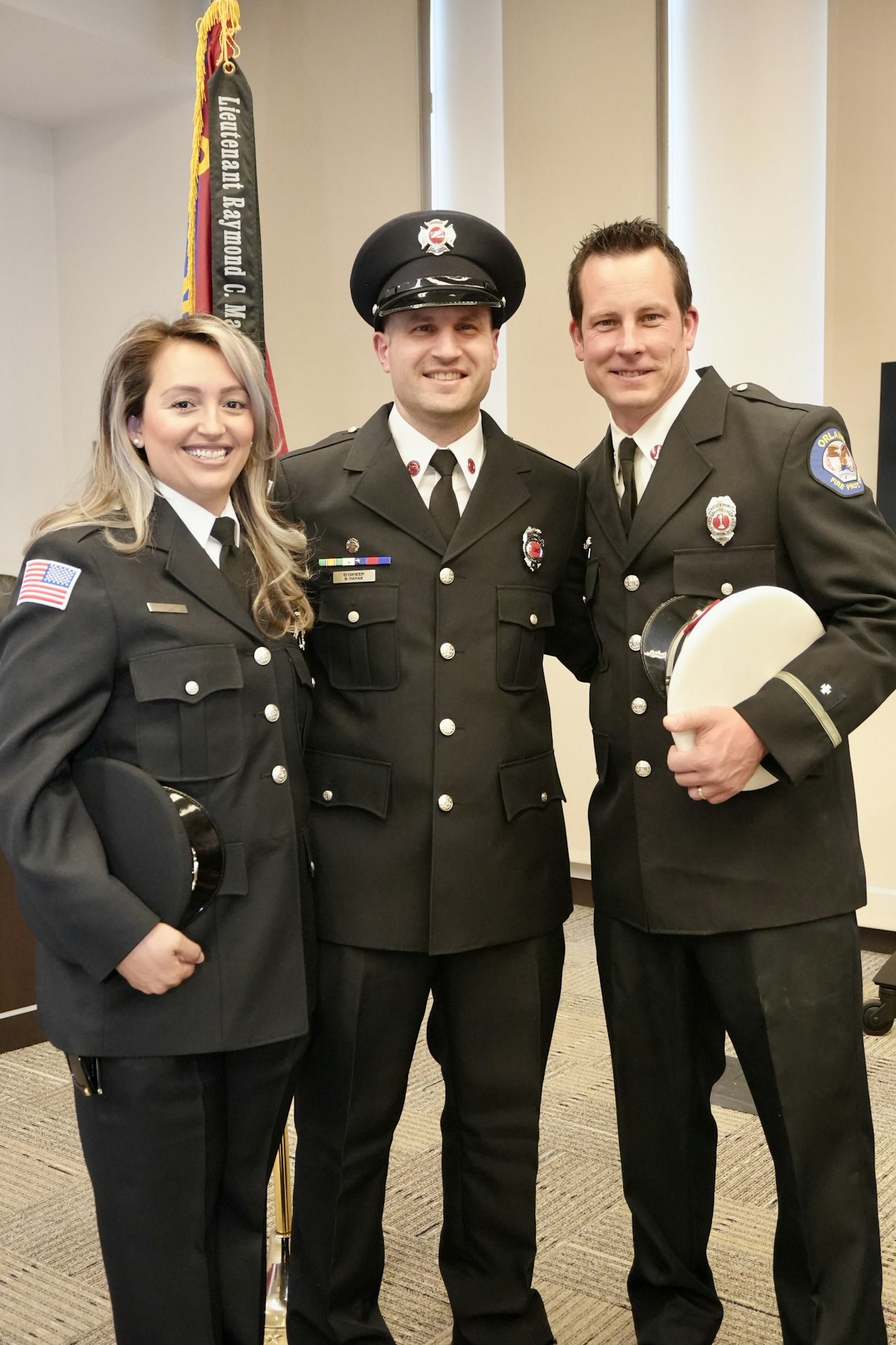 Three people in uniform, smiling, standing together indoors with a banner in the background.