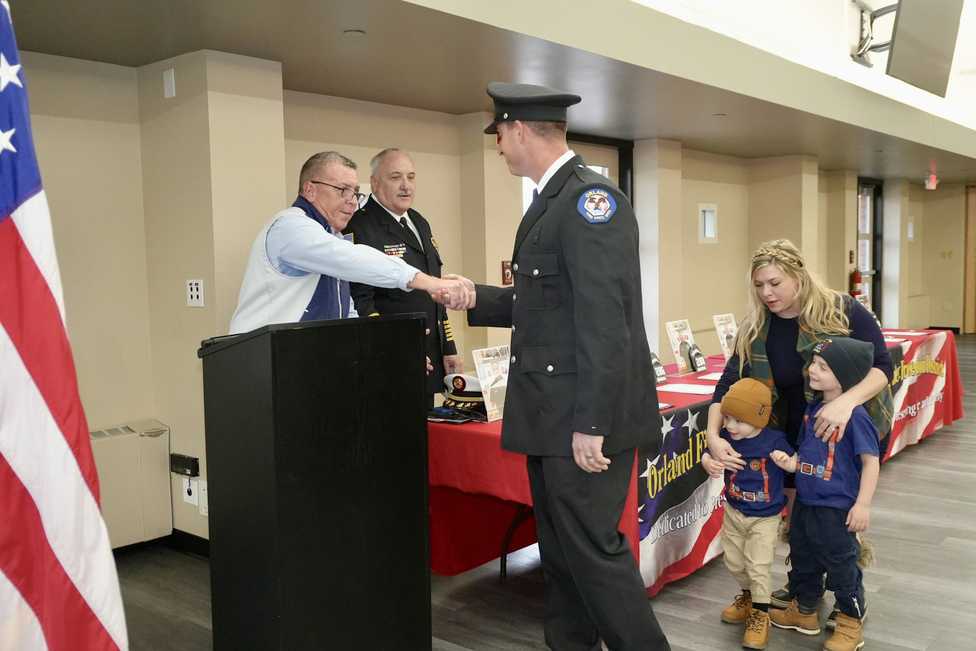A man in uniform shakes hands with another man beside a podium, while a woman with two children stands nearby. An American flag is visible.