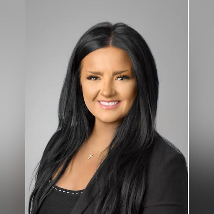 A person with long dark hair, wearing a black suit and a cross necklace, smiling against a gray background.