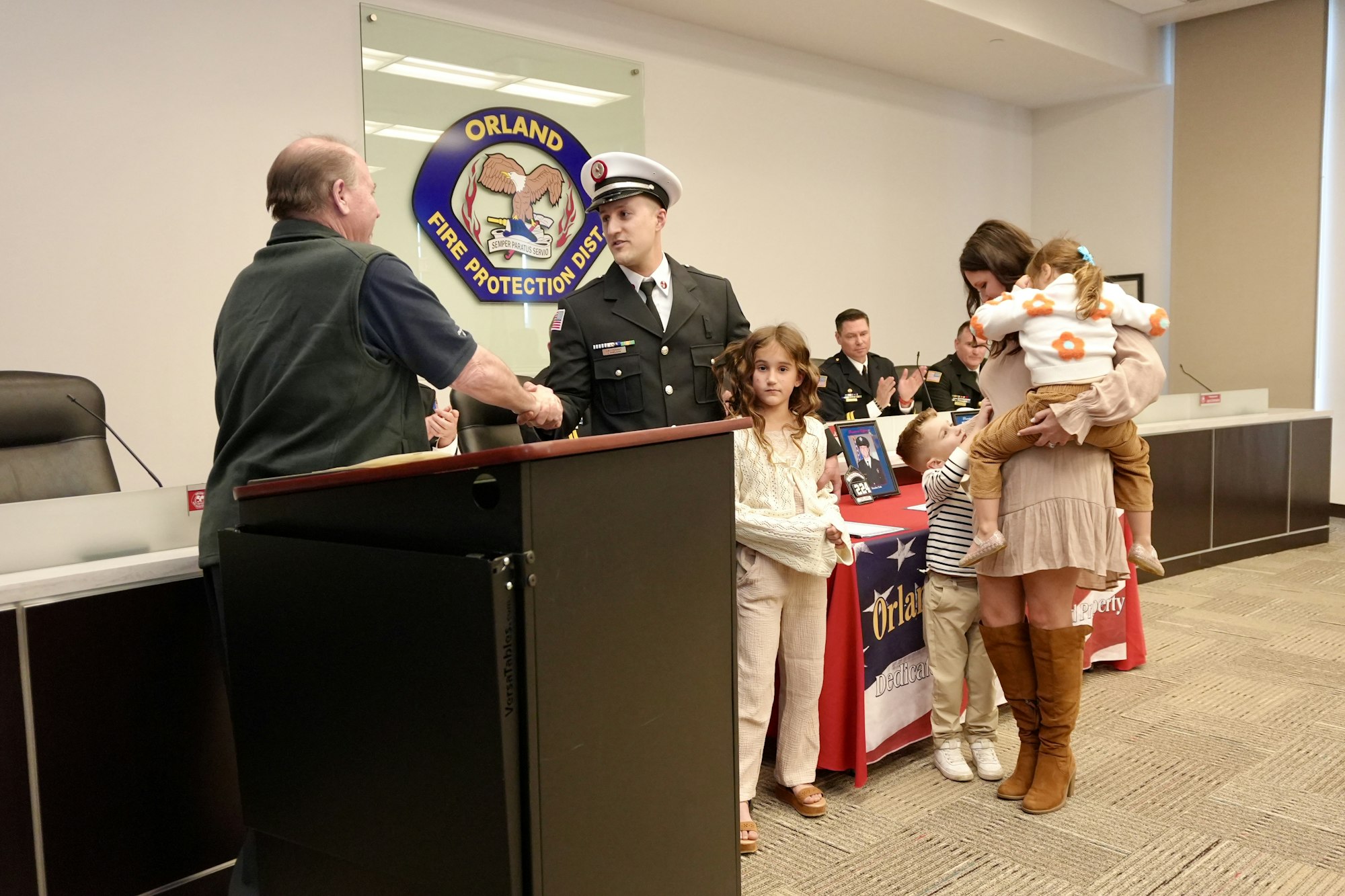 A man in a uniform shakes hands; a family stands nearby. Background shows the Orland Fire Protection District logo.