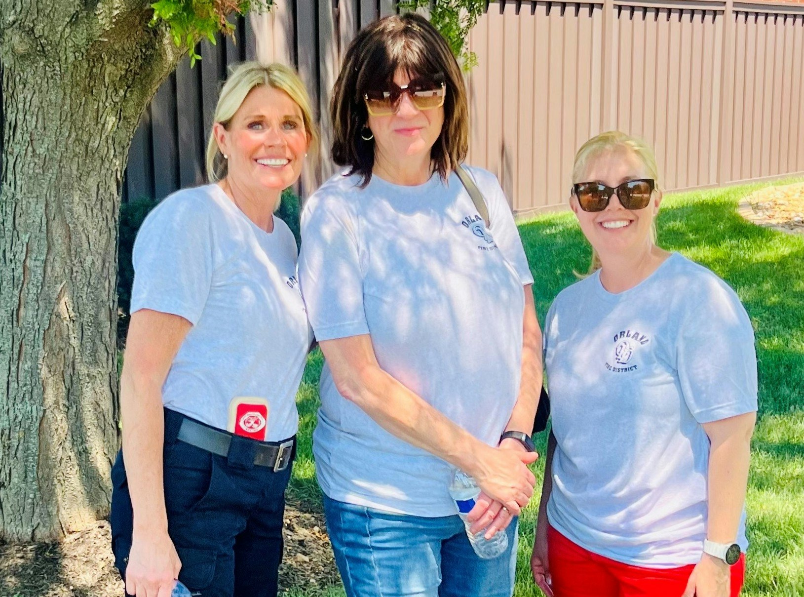 Three women wearing sunglasses and matching gray T-shirts stand outdoors near a tree.