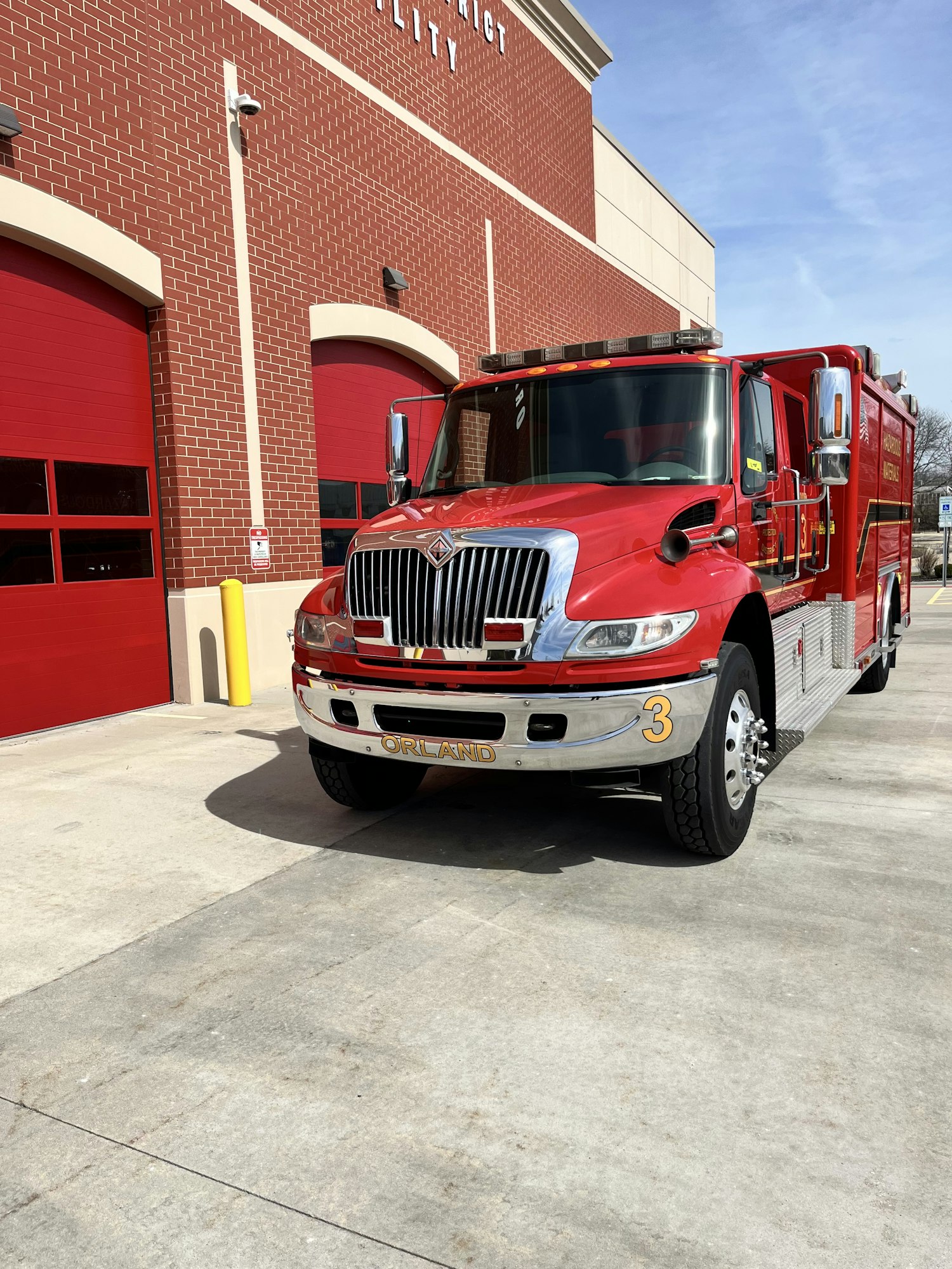 A red fire truck parked in front of a fire station with brick walls and red doors. Clear sky overhead.