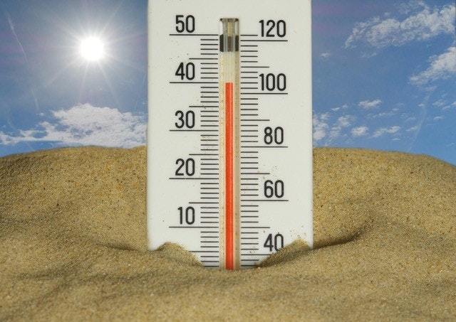 Thermometer in sand under a bright sun and blue sky, showing high temperature.