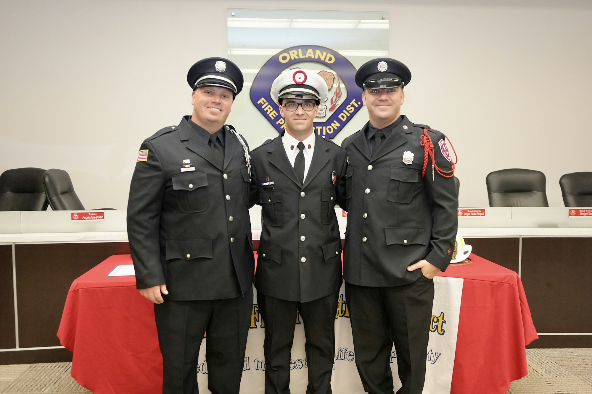 Three uniformed firefighters stand together, smiling inside a meeting room with the Orland Fire Protection District logo in the background.
