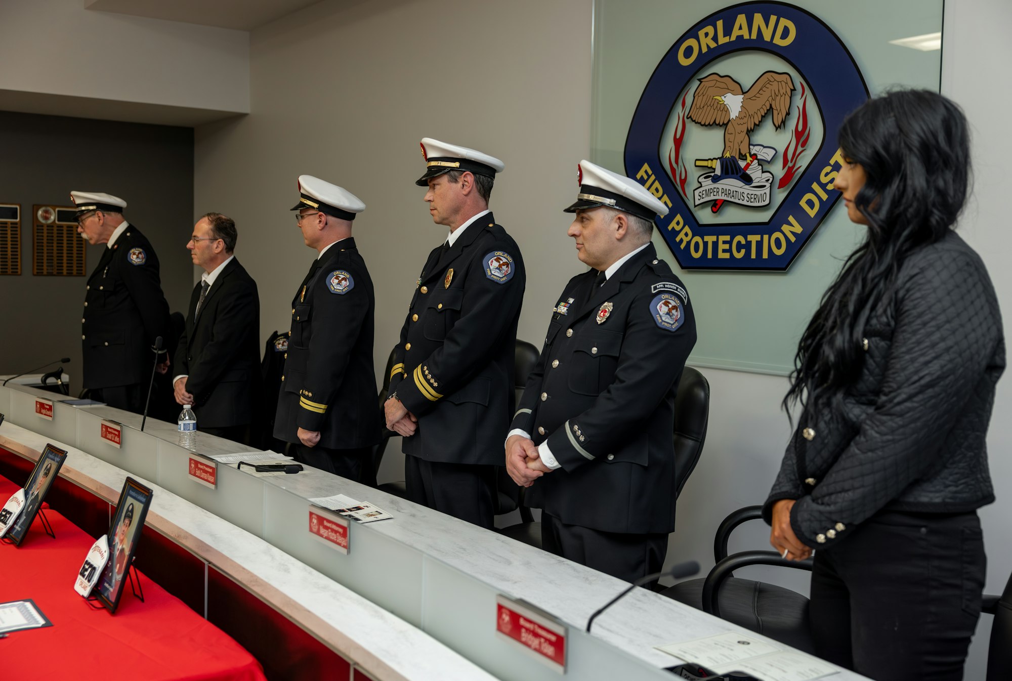 A group of uniformed fire officials stands at a ceremony with plaques, while a woman observes. There's an Orland Fire Protection District logo.