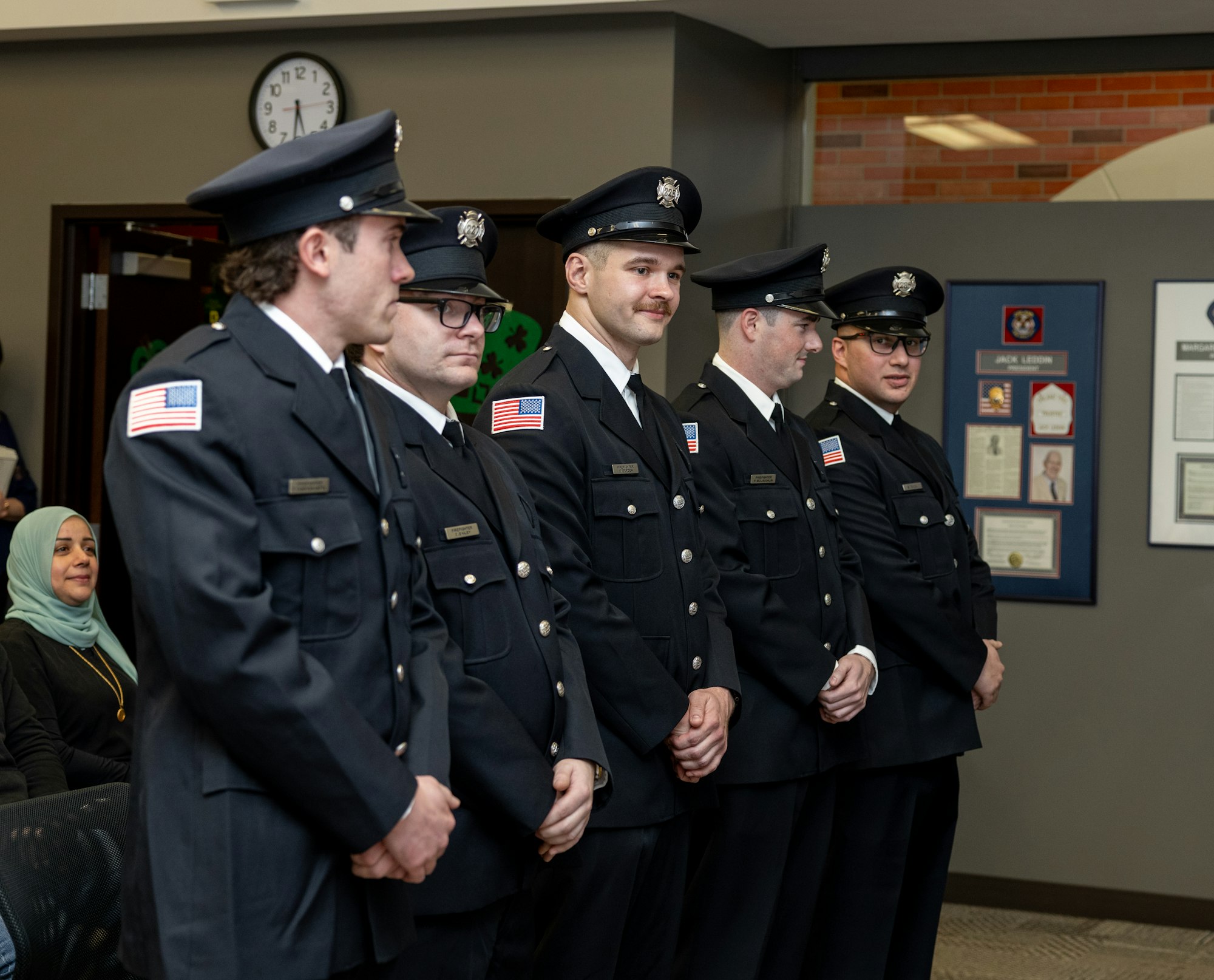 A line of uniformed individuals, likely firefighters or police officers, stands in a ceremony, with an audience member visible in the background.