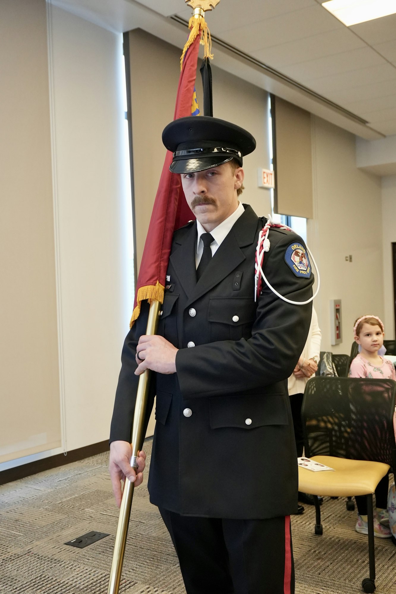 A uniformed person holding a flag stands indoors with a young child in the background.