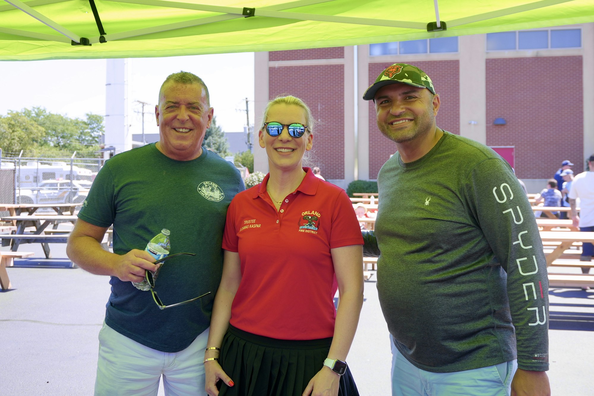 Three people smiling under a green tent, outdoors, near benches and buildings.