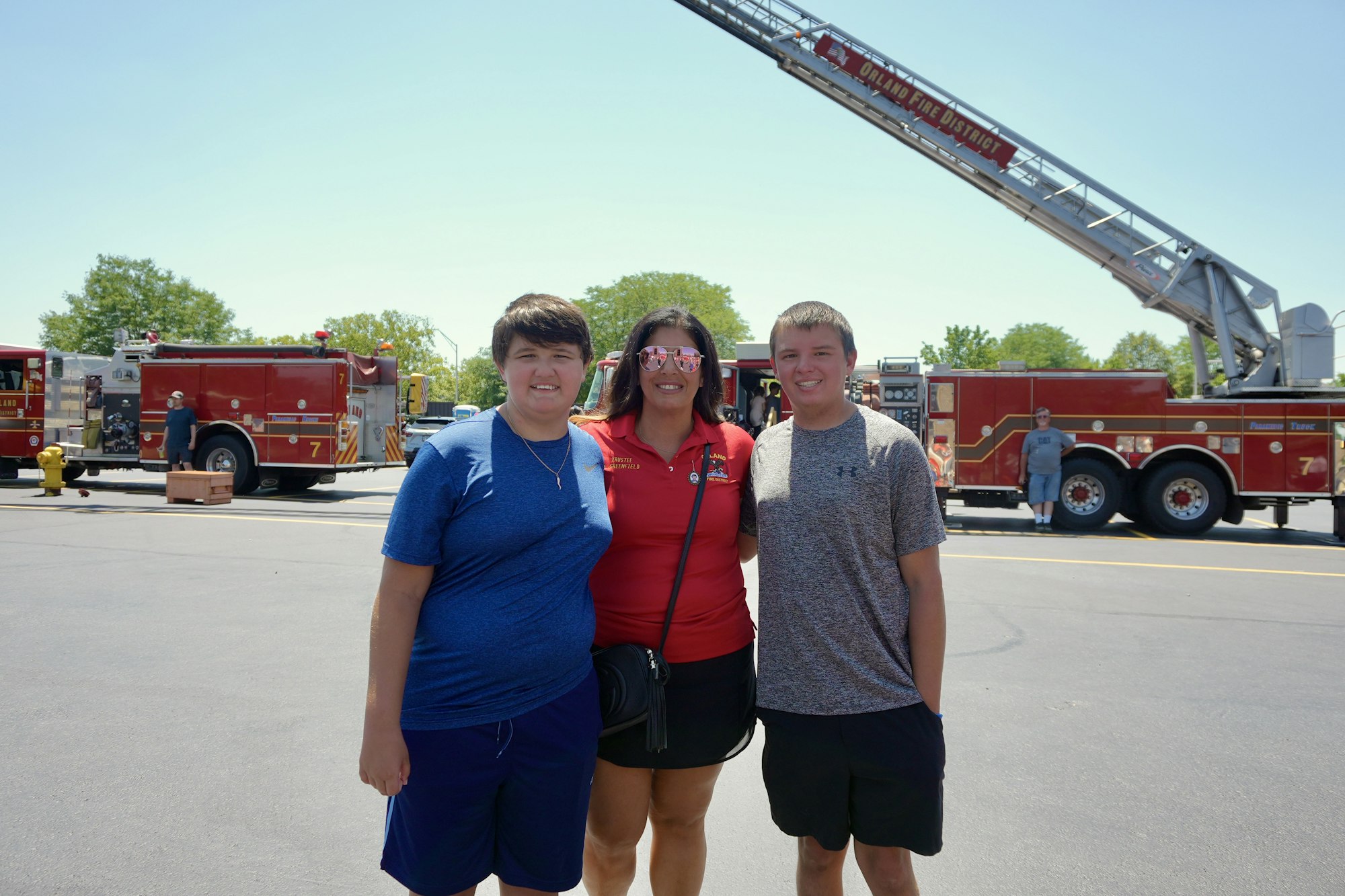 Three people posing happily in front of fire trucks with extended ladders in a parking lot.