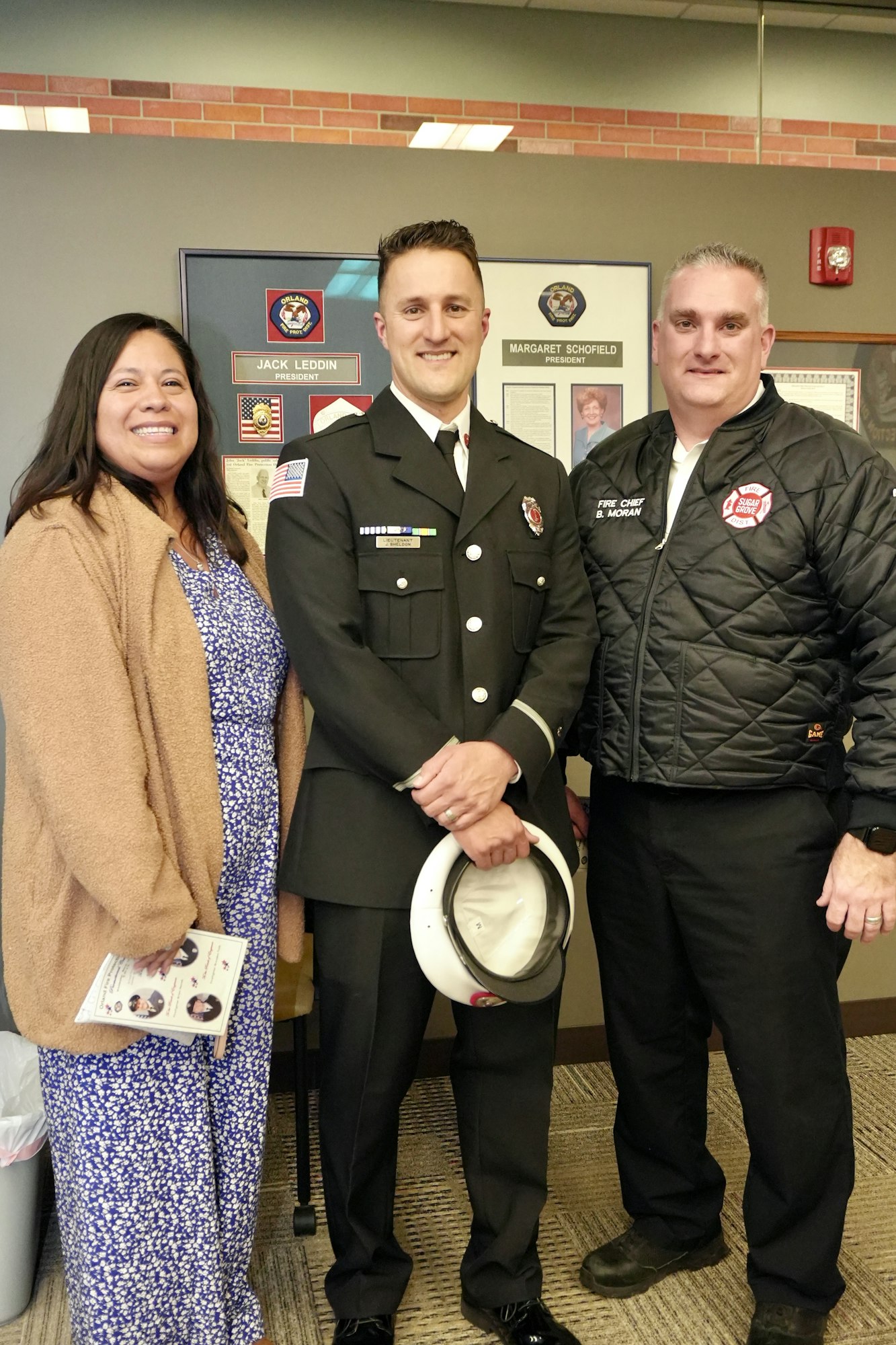 Three people, one in a fire uniform, smile while standing indoors.