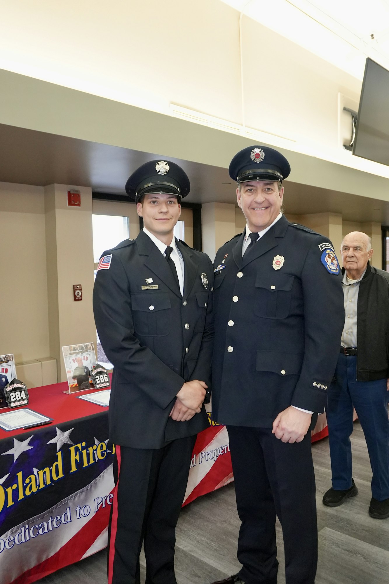 Two smiling firefighters in uniform pose indoors at an event, with a table and an older man in the background.