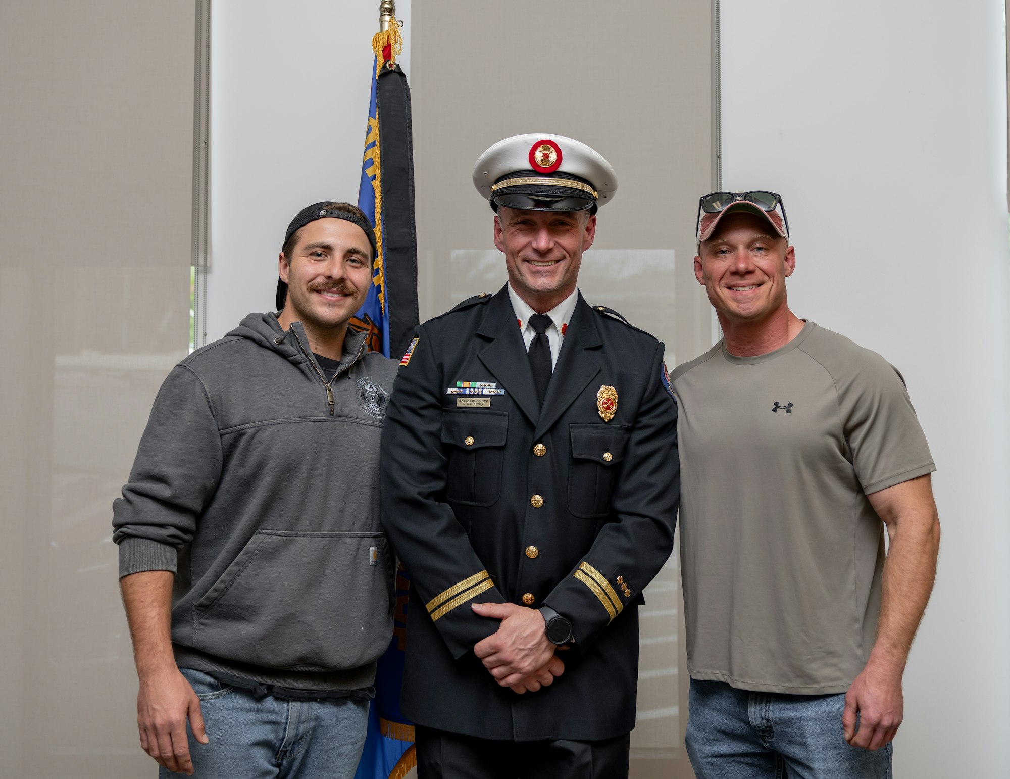 Three men pose for a photo. One is in a formal uniform, while the others are in casual clothing, smiling together.