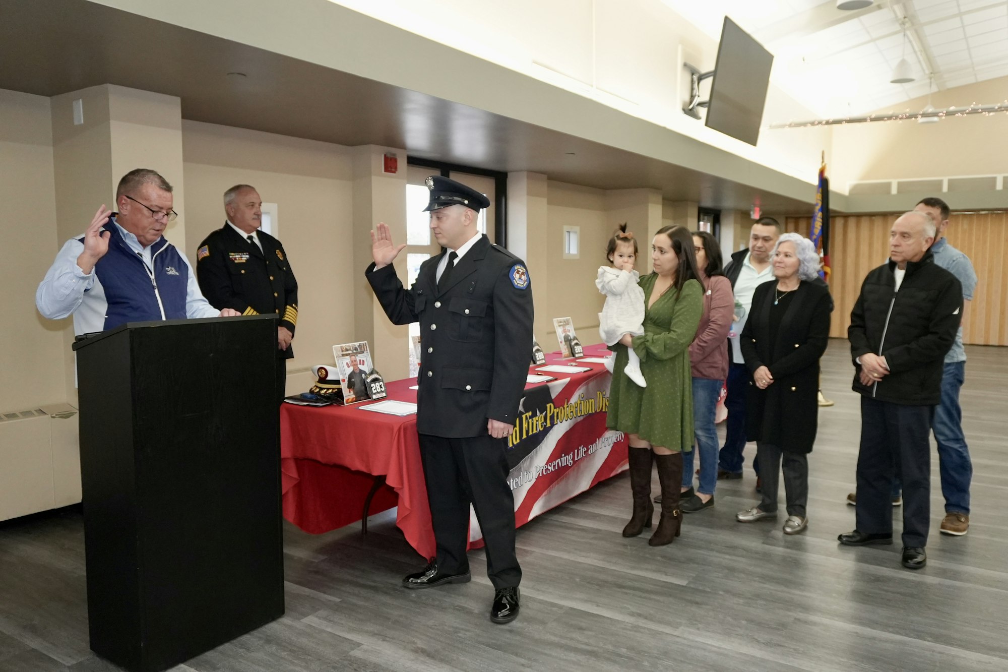 A firefighter is being sworn in, with family and officials present. A child is held by a woman, and a banner is visible on a table.