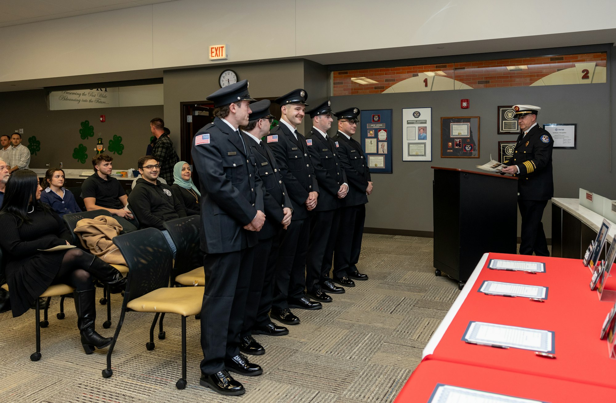 A formal ceremony with uniformed officers standing in line, while a speaker addresses an audience in a community space.