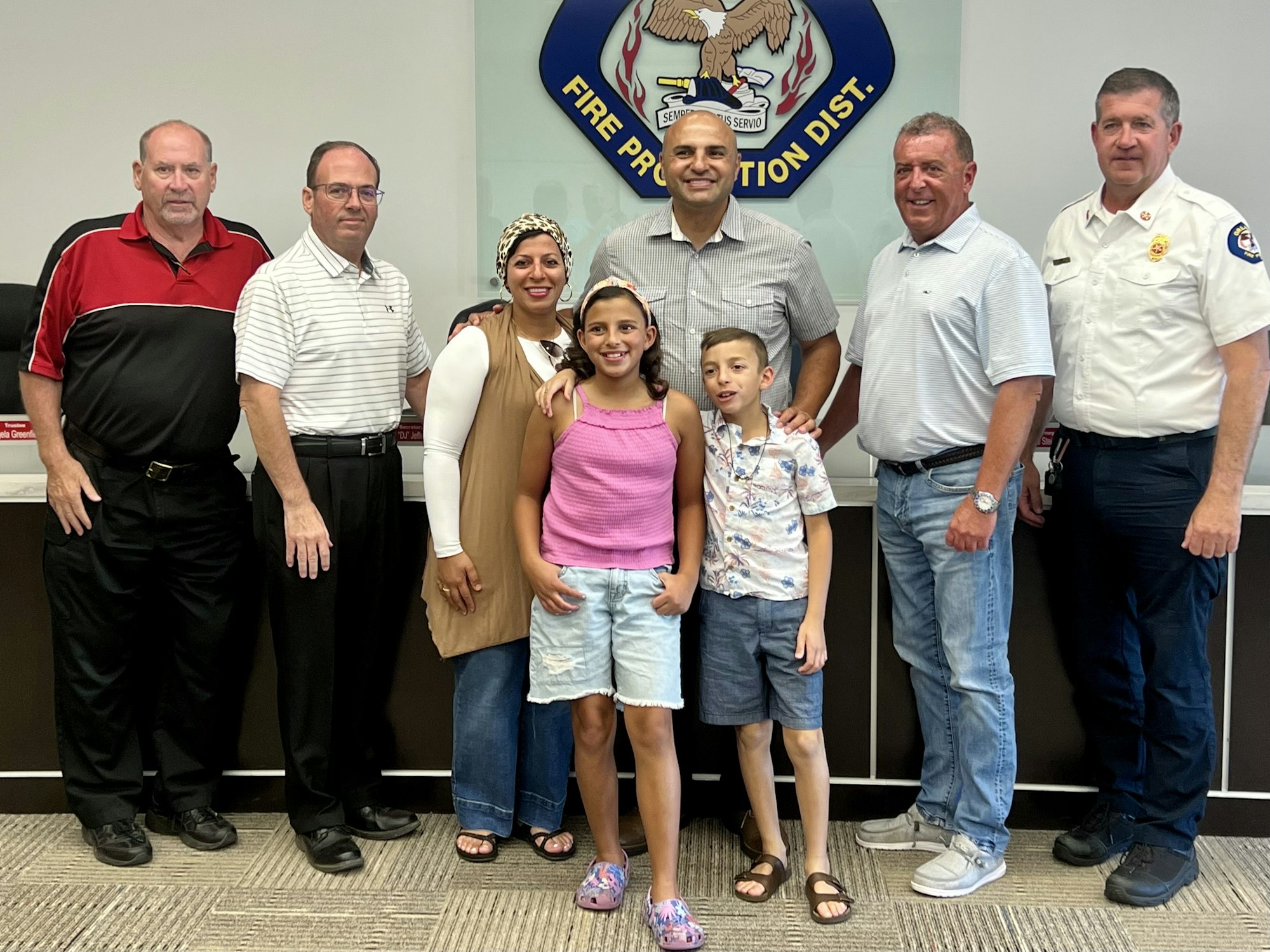 A group of adults and two children pose indoors in front of a "Fire Protection Dist." sign.
