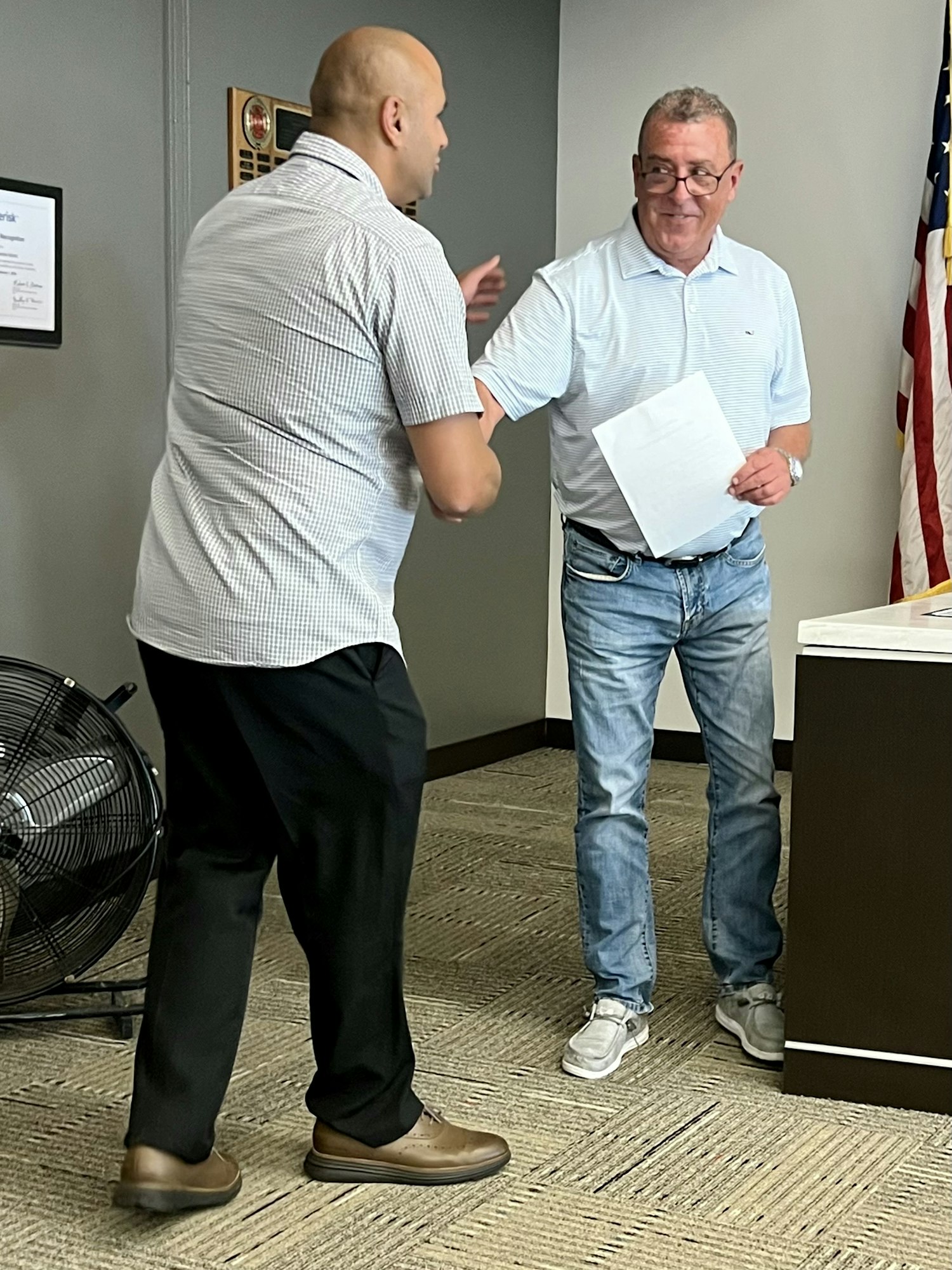 Two people in an office setting, one holding a paper, appear to be shaking hands. An American flag is visible.
