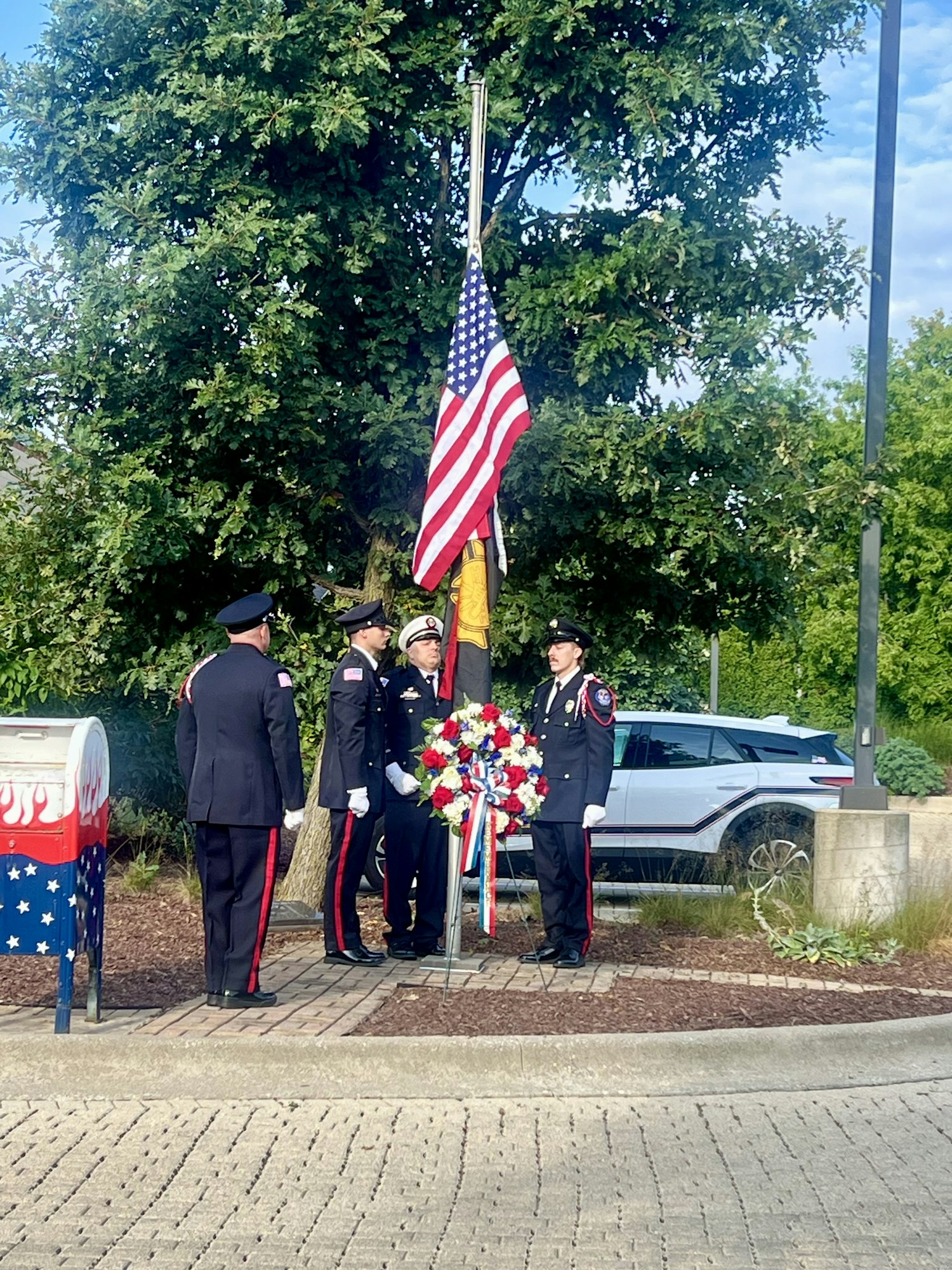 Uniformed personnel in a ceremony with a U.S. flag and a wreath, standing near a decorated mailbox.