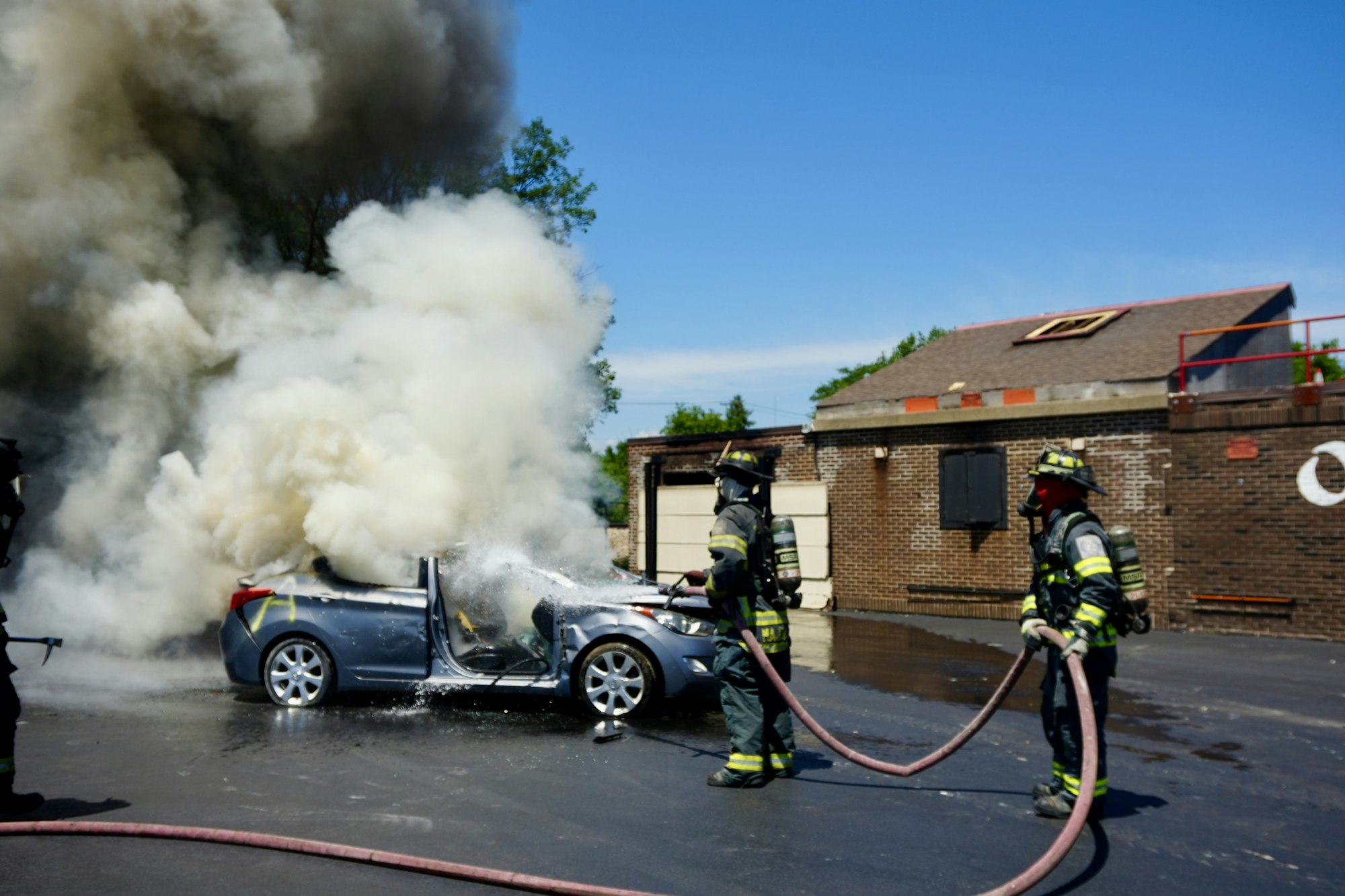 Firefighters extinguishing a car fire with hoses, surrounded by heavy smoke.
