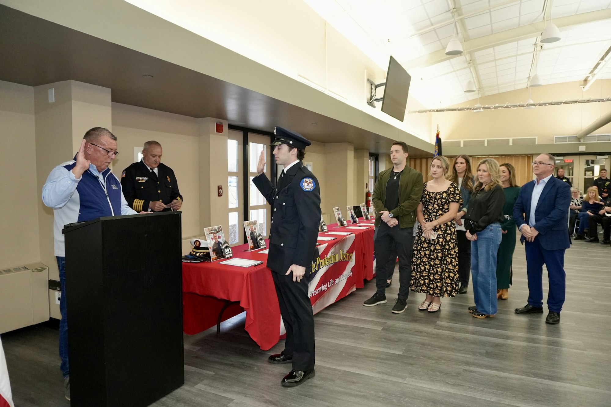 A man in uniform is sworn in at a ceremony with people watching. There’s a red table with photos and items on it.