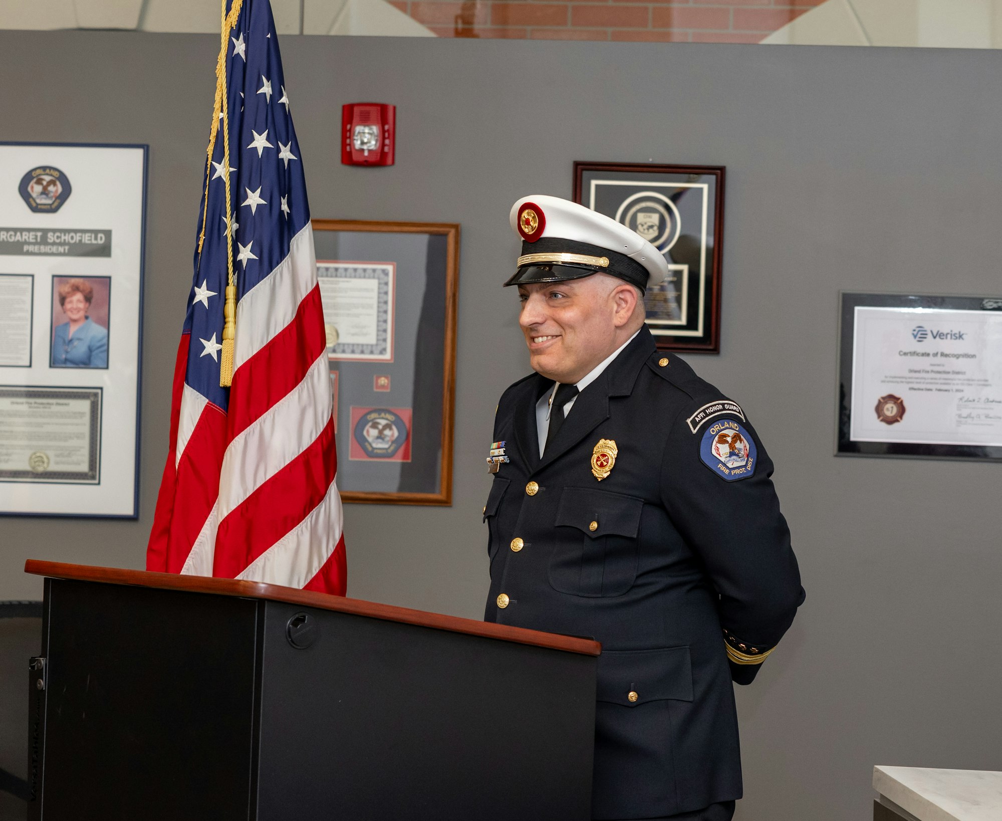A smiling officer in uniform stands at a podium, with an American flag beside him and recognition certificates on the wall.