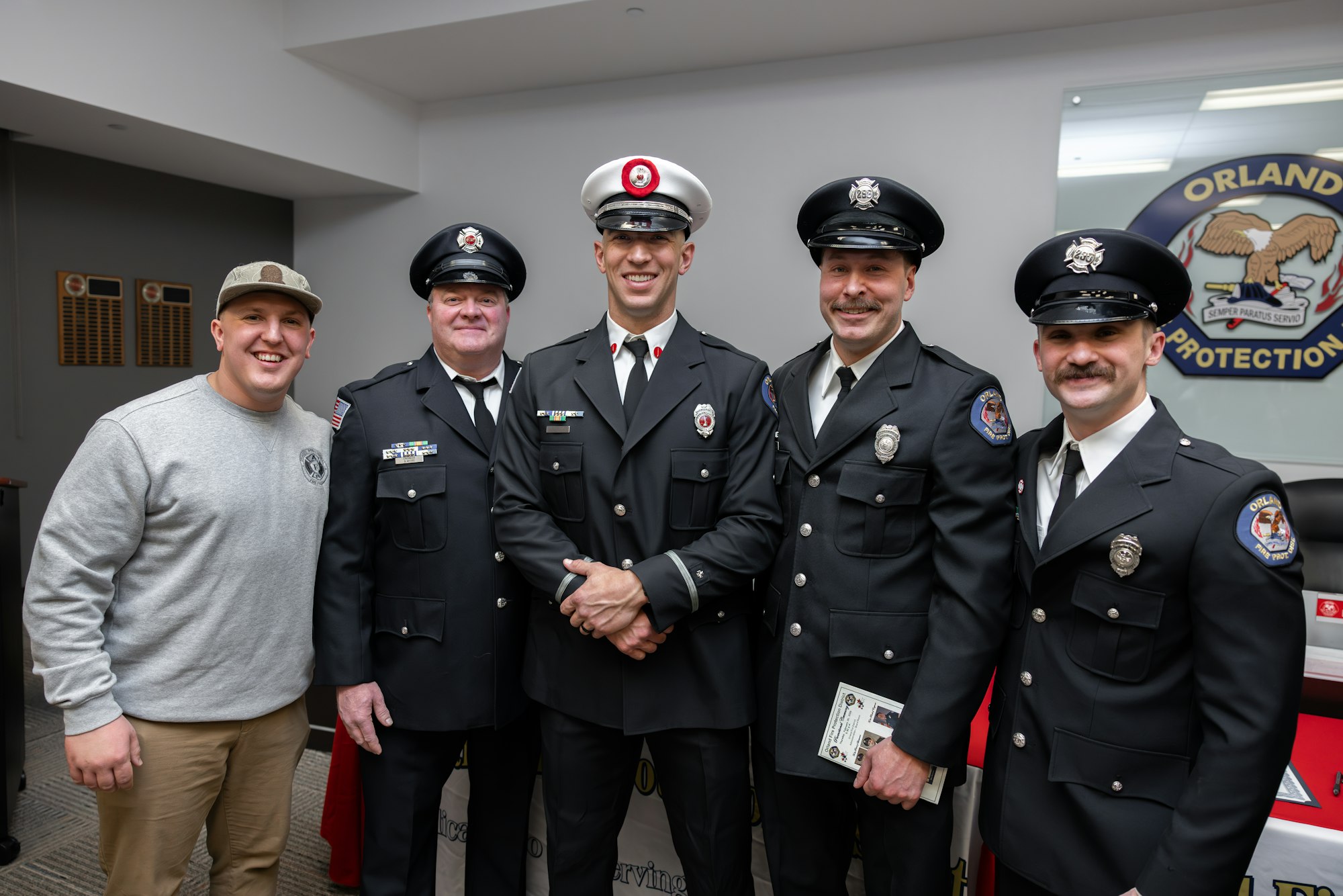 A group of five men in uniform, including firefighters and police, posing together in a room with an official emblem visible.