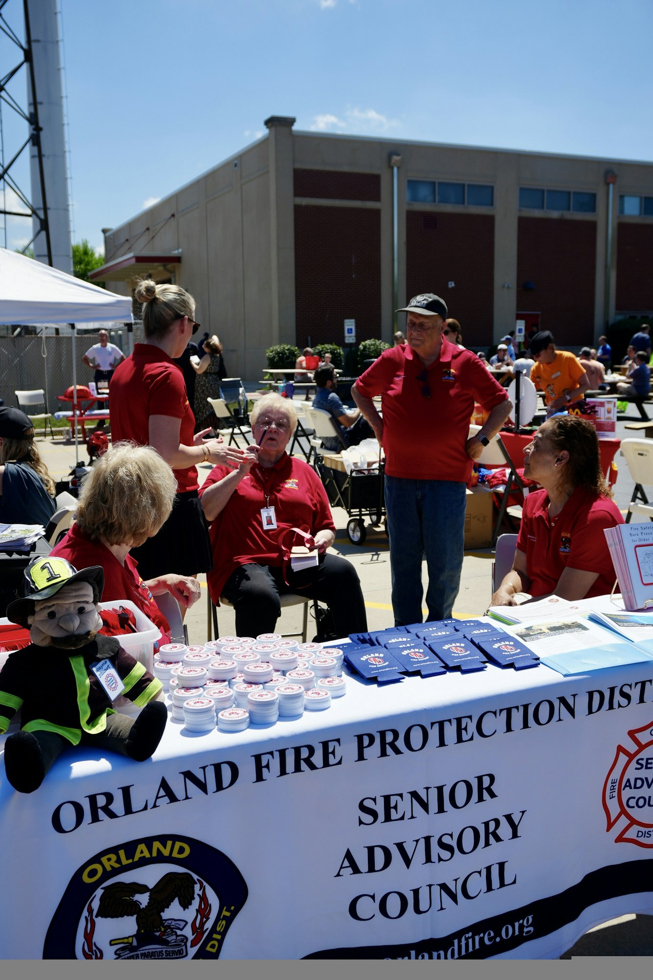 People in red shirts at an Orland Fire Protection District event, with a table displaying items like brochures and a firefighter doll.