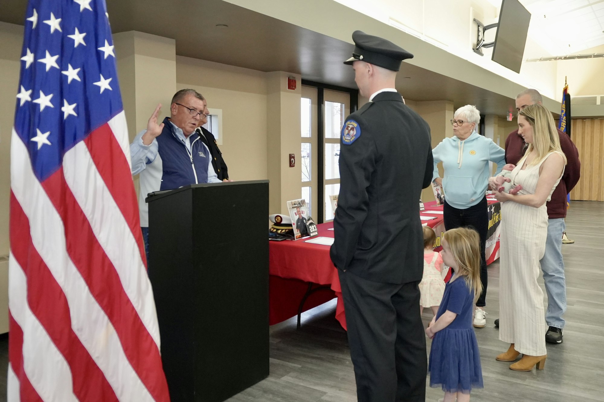 A person is being sworn in by a podium, facing another in uniform, with family and a flag nearby.