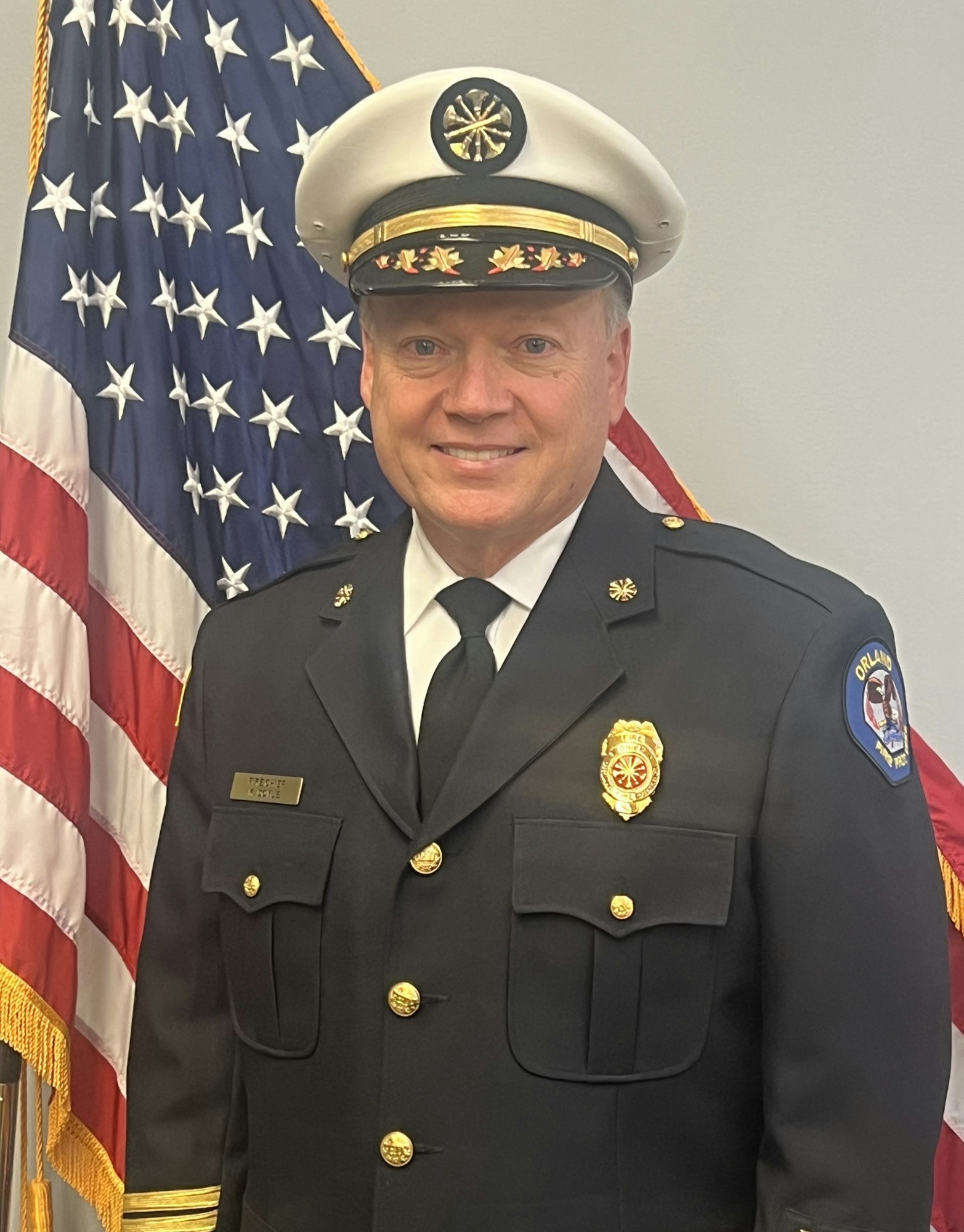 A person in a formal police or fire chief uniform stands in front of an American flag, smiling for the photo.
