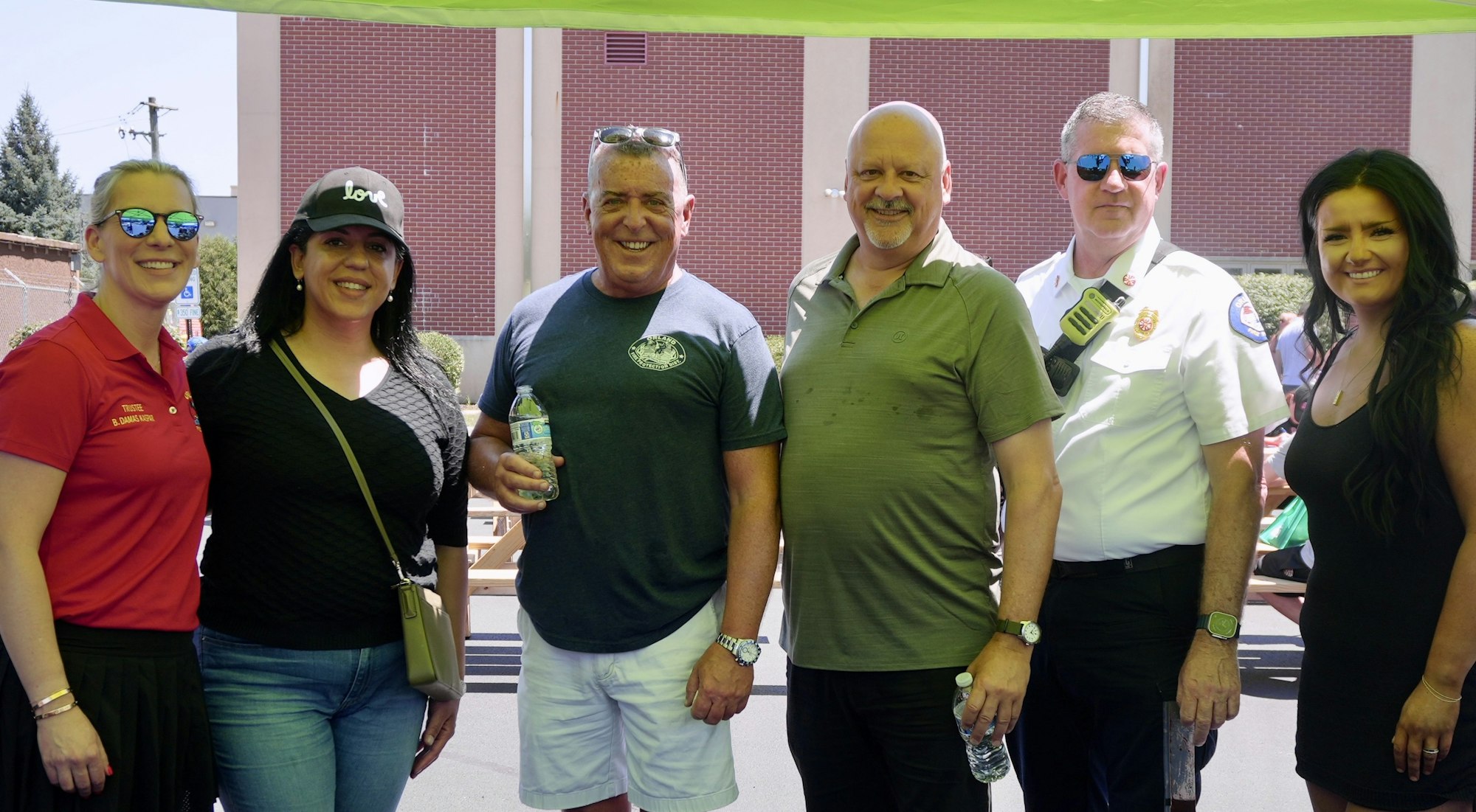 A group of six people posing outdoors, some holding water bottles, with a brick building in the background.