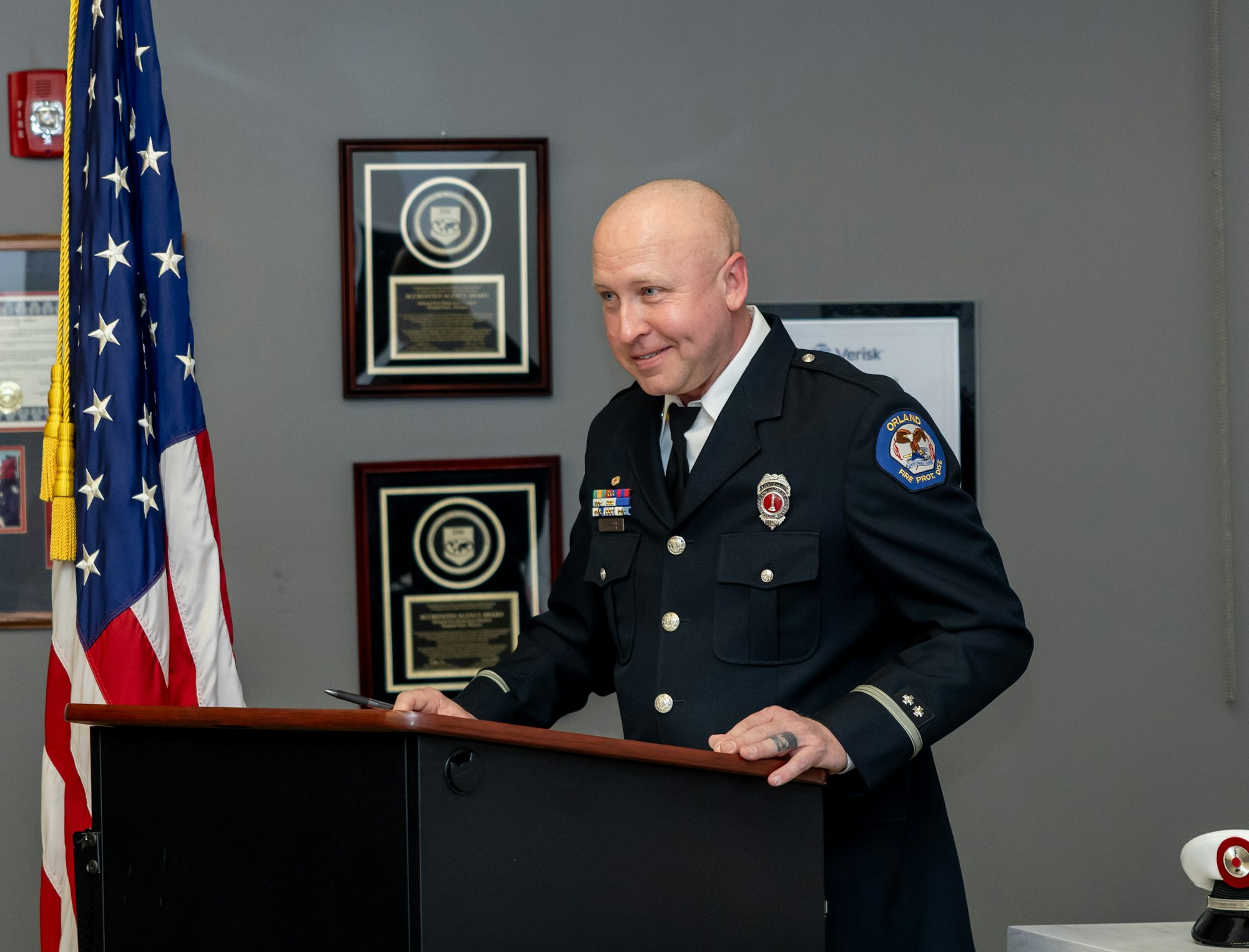 The image shows a uniformed officer at a podium, smiling, with an American flag in the background and plaques on the wall.