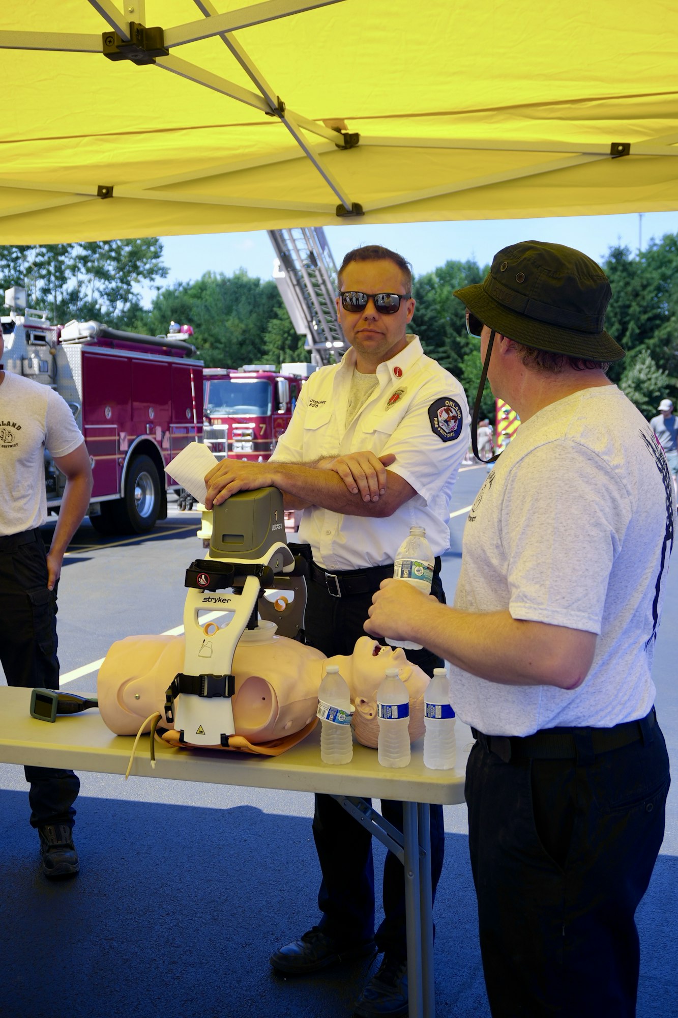 People under a yellow tent with CPR training equipment and a mannequin on a table; fire trucks are in the background.