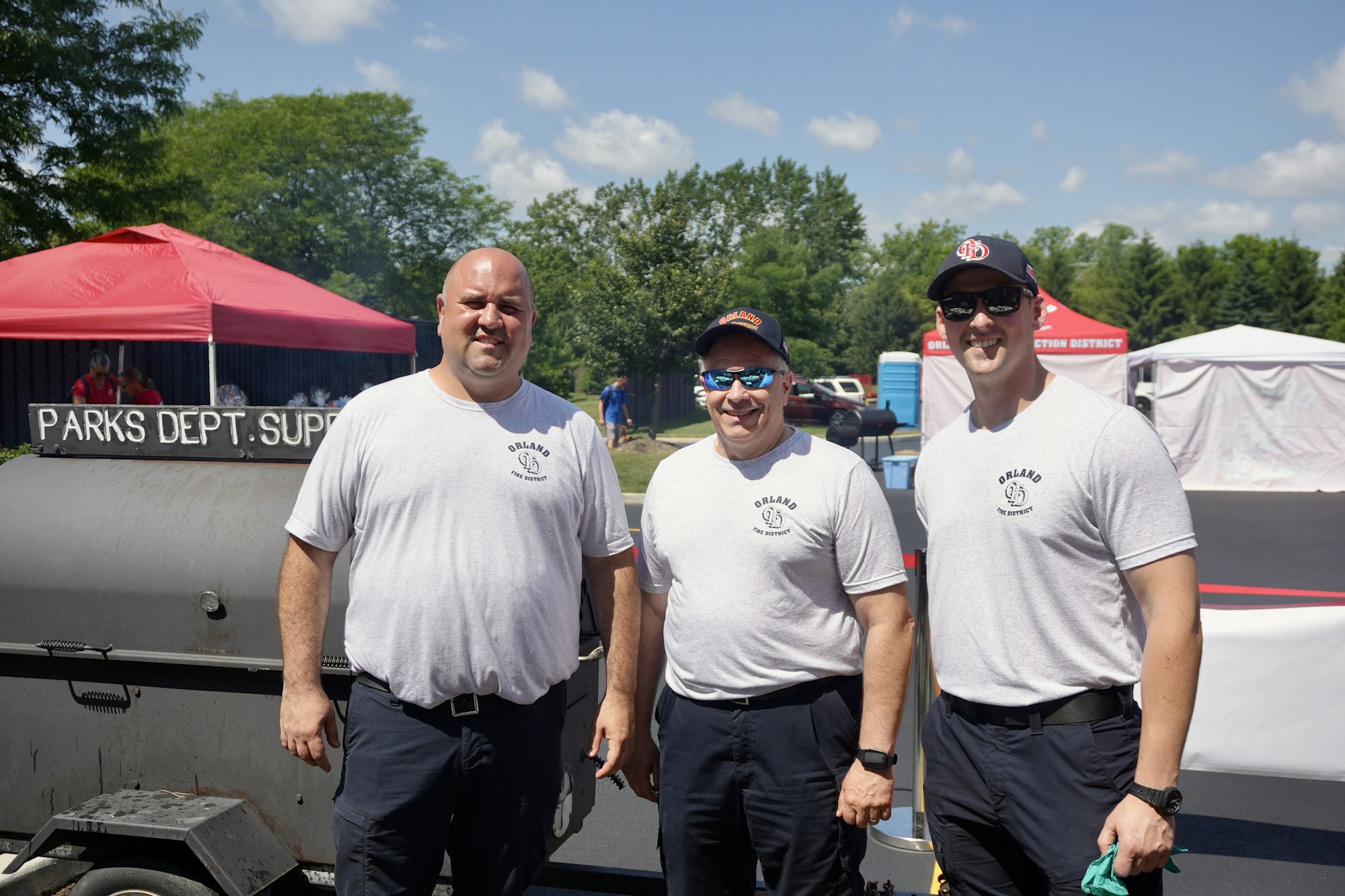 Three people in matching shirts stand in front of a grill or smoker, with event tents and trees in the background.