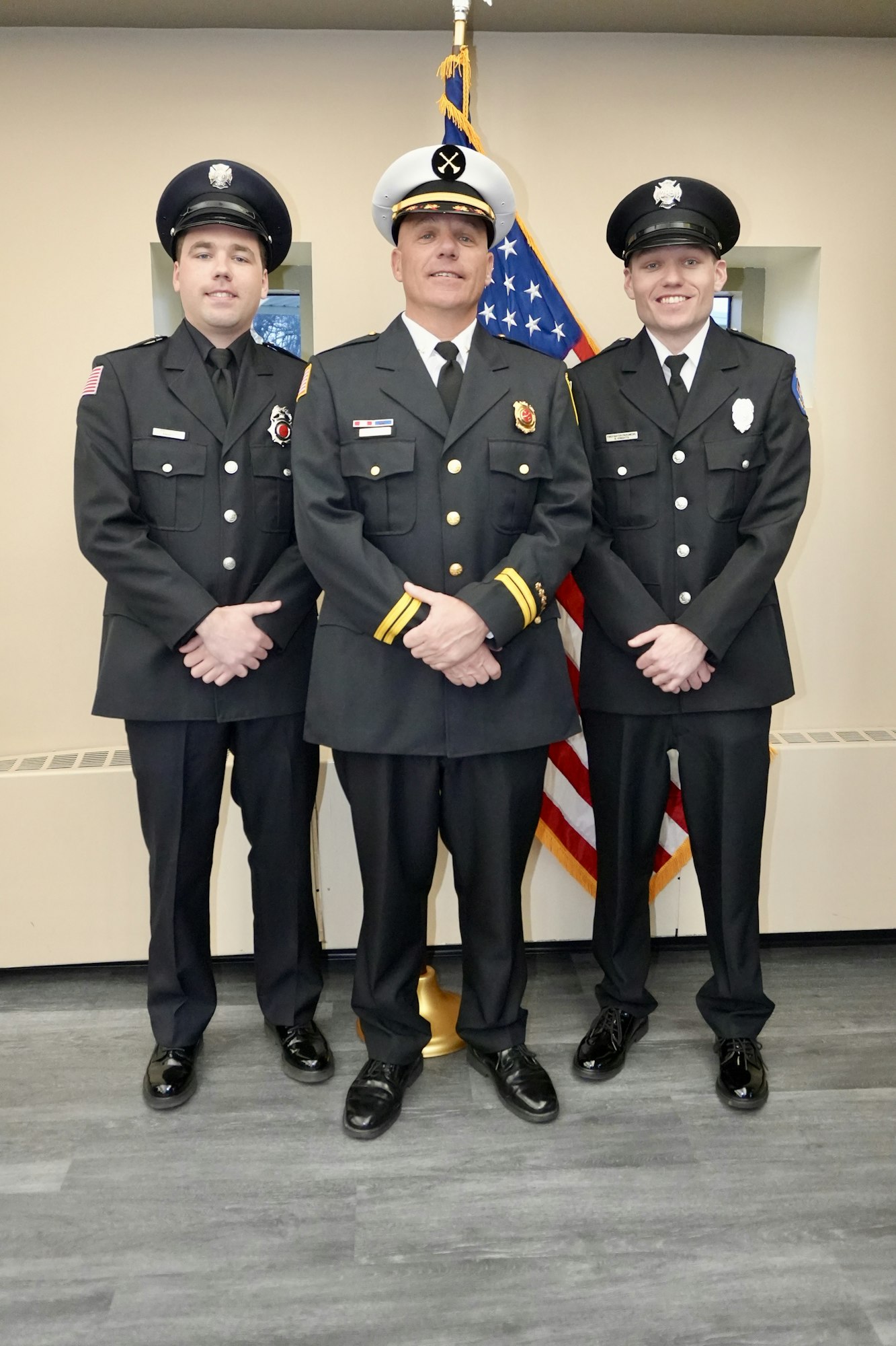 Three firefighters in uniforms stand in front of a U.S. flag.