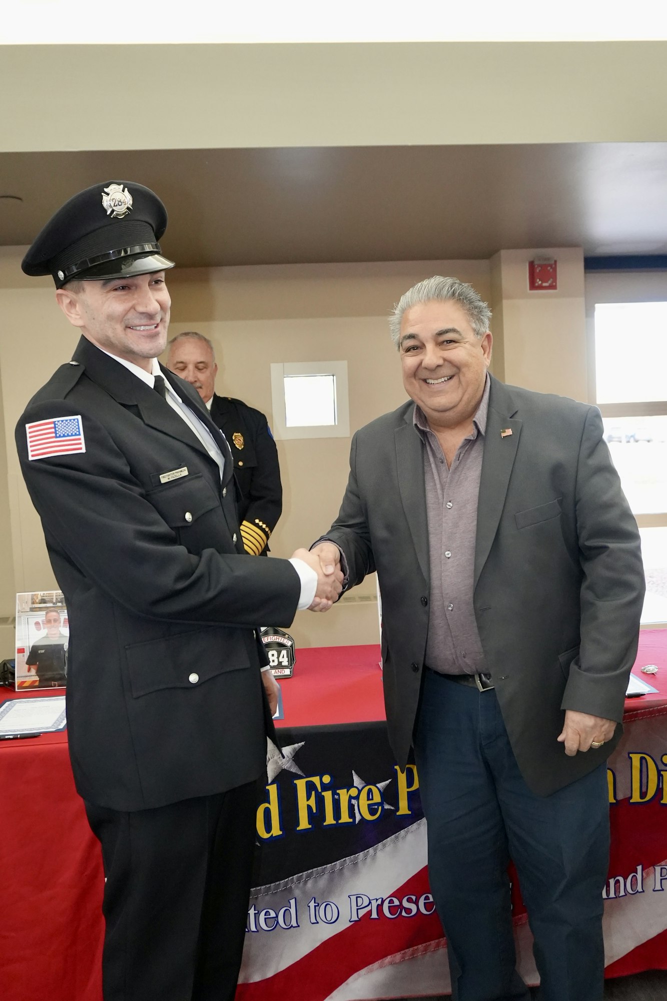 A firefighter and another man in business attire shake hands at an event, smiling, with a banner in the background.