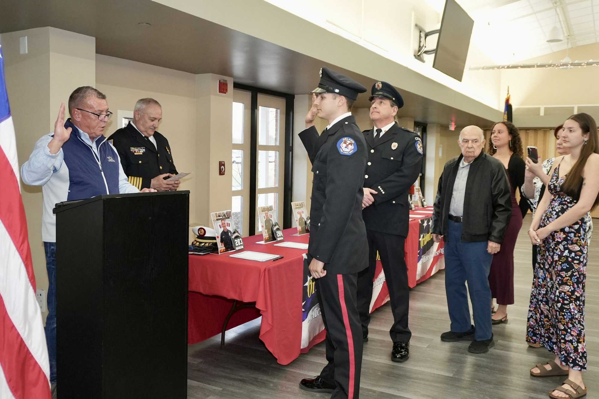 A man in uniform salutes during a ceremony with officials and onlookers nearby, next to a table with photos and memorabilia.