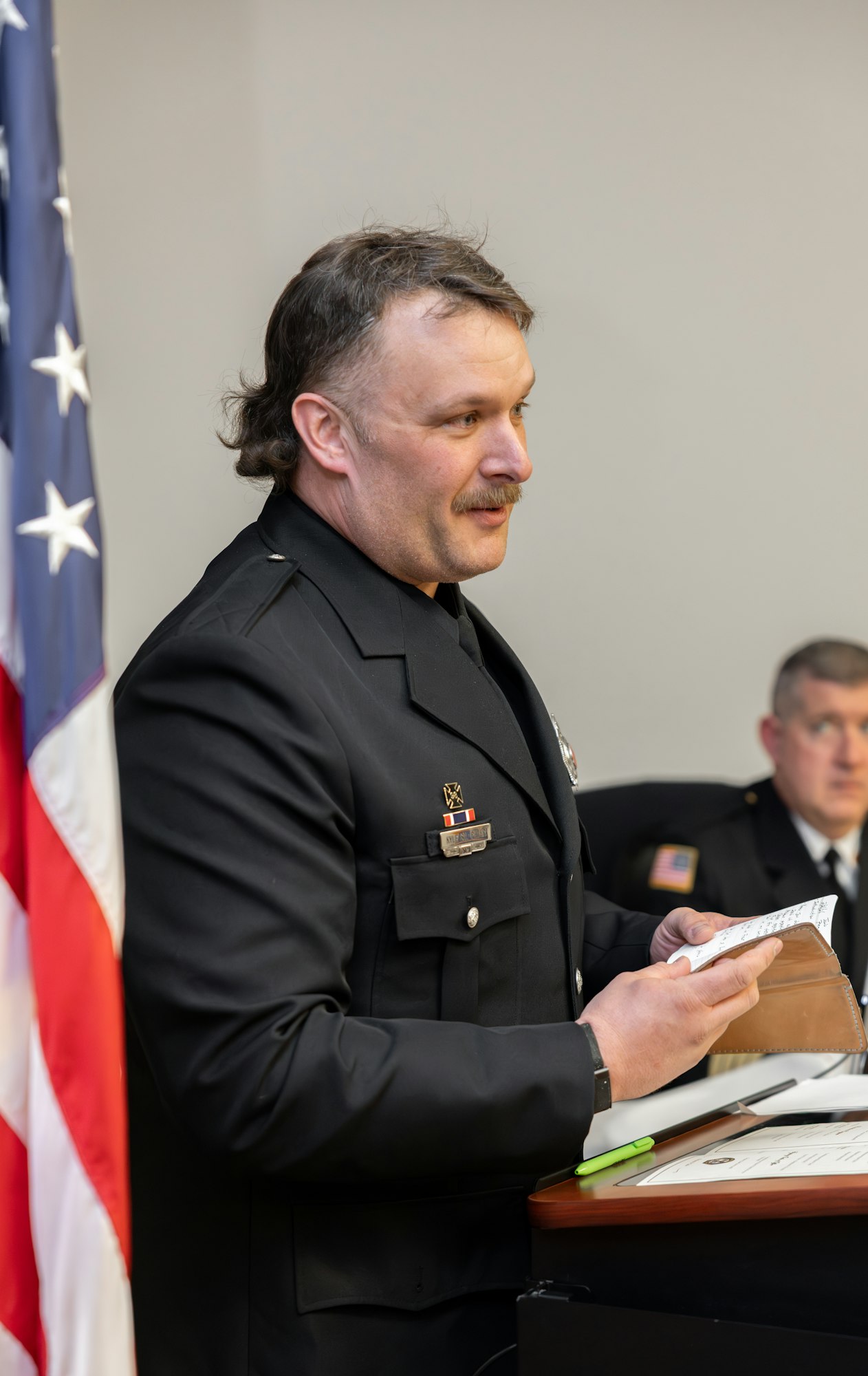 A man in a police uniform is speaking at a podium, with an American flag in the background and another officer seated nearby.